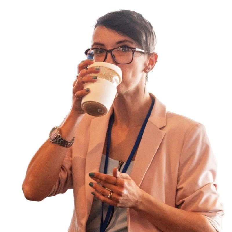 A woman with short dark hair, glasses, and earrings, wearing a pink blazer and a lanyard, is drinking coffee or tea from a paper cup.