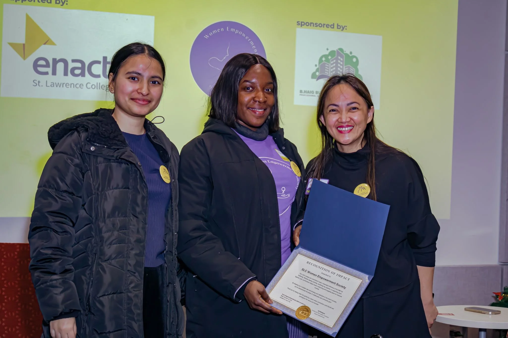 Three women smiling and standing together, one holding a certificate, in front of a yellow background with logos and text, at an award or recognition event.