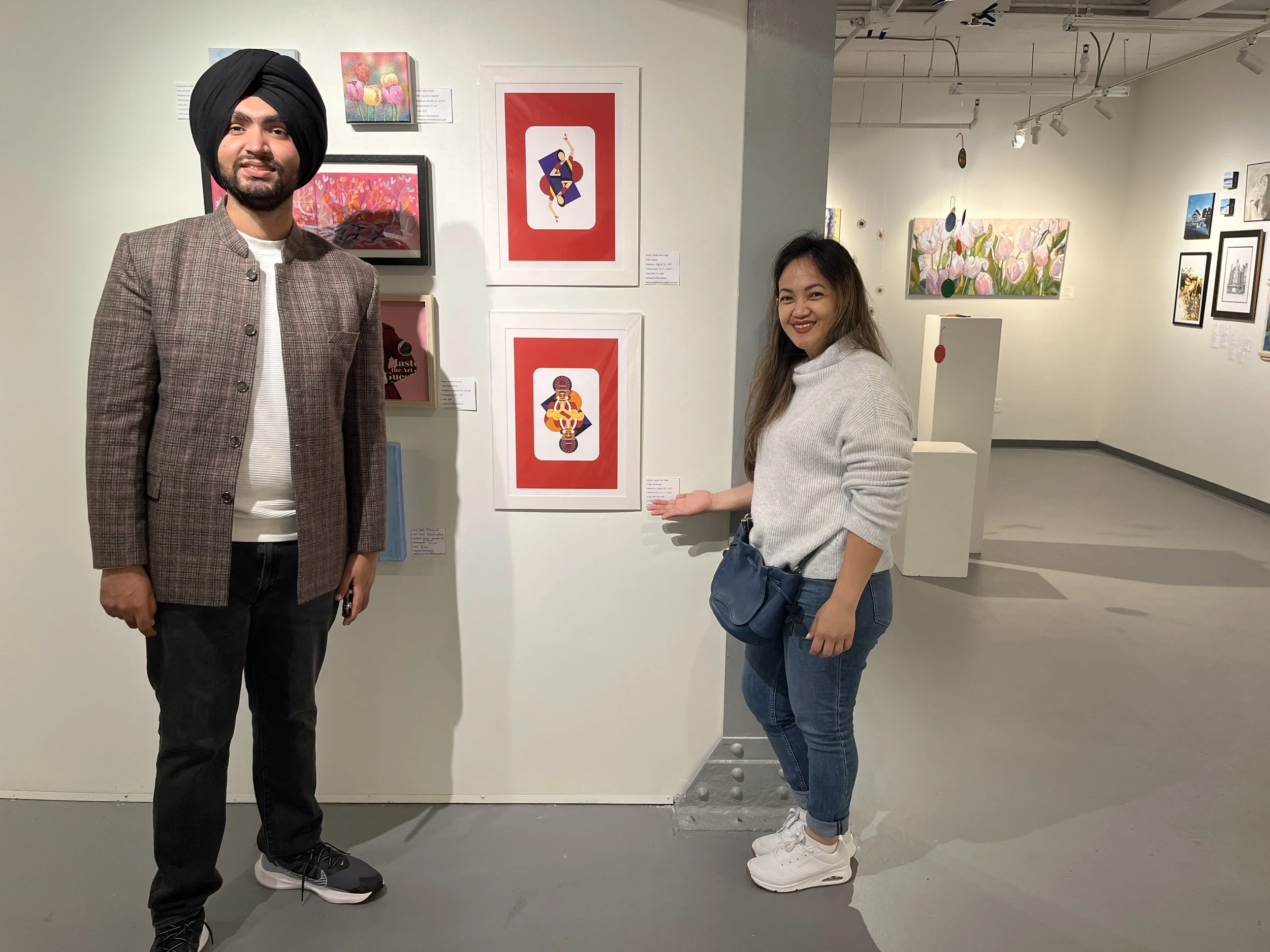A man and woman standing in an art gallery next to colorful artwork on the wall. The man is wearing a black turban, blazer, white shirt, and black pants. The woman is wearing a light grey sweater and blue jeans with white sneakers, smiling and pointing towards the artwork.