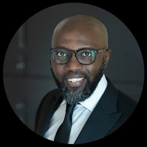 Portrait of a man with glasses, a beard, wearing a black suit and tie, smiling, with a dark background.