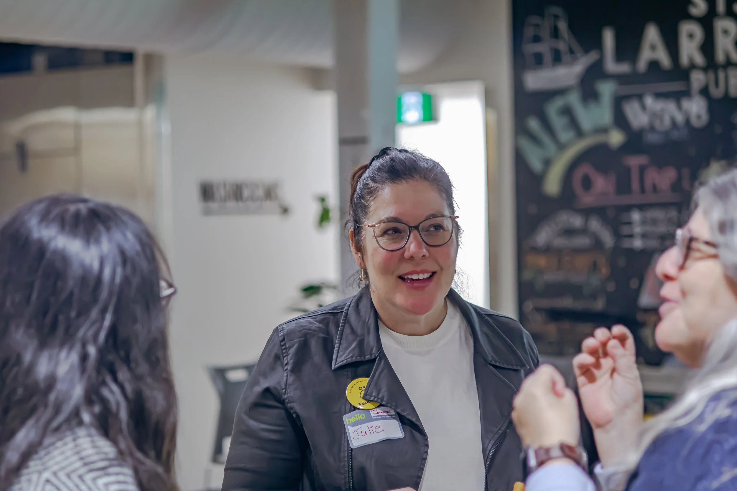 A woman with glasses, wearing a black jacket and a name tag that reads 'Julie,' is smiling and talking with two other women in an indoor setting.
