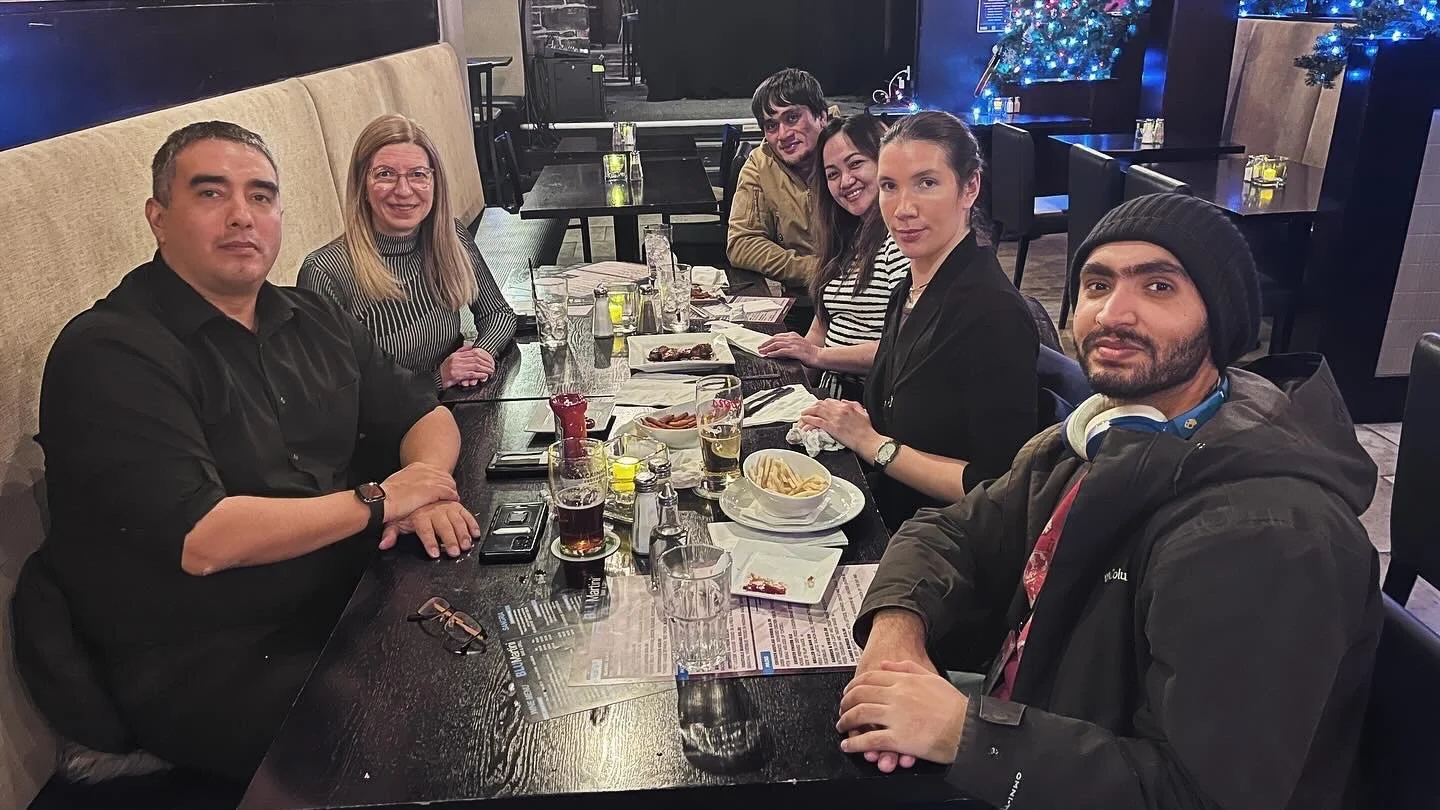 Group of six people sitting at a long table in a dimly lit restaurant with holiday decorations and blue lights in the background.