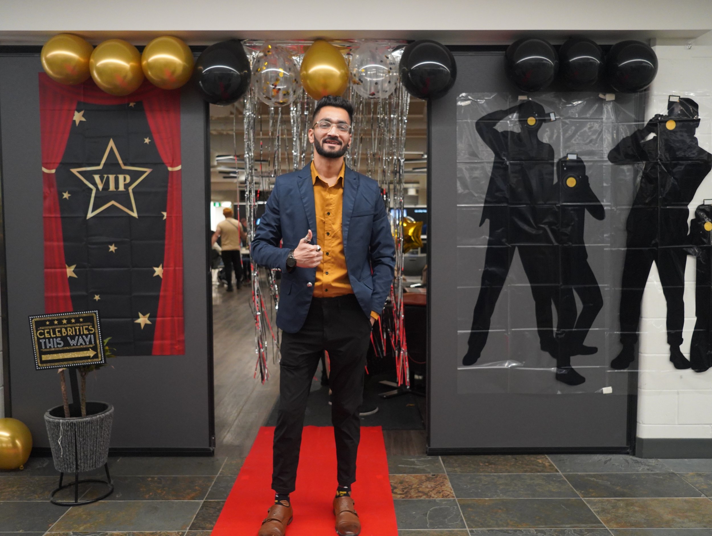Man in blue blazer and yellow shirt standing on red carpet at celebration with VIP sign, balloons, and silhouette decorations.