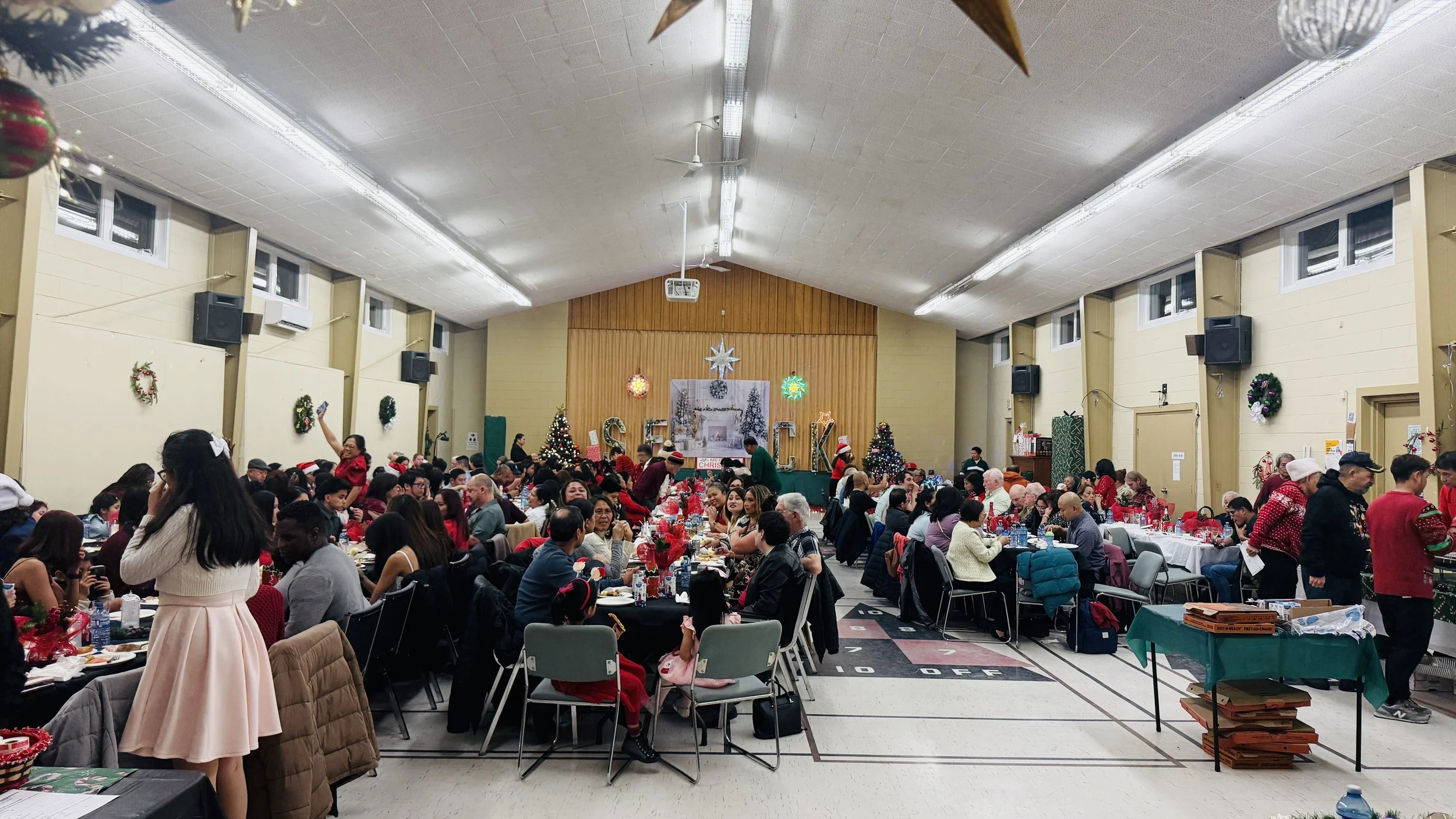 A large indoor Christmas celebration with many people sitting at decorated tables, some wearing Santa hats, in a gymnasium with holiday decorations, including Christmas trees, wreaths, and a decorated stage with festive banners and ornaments.