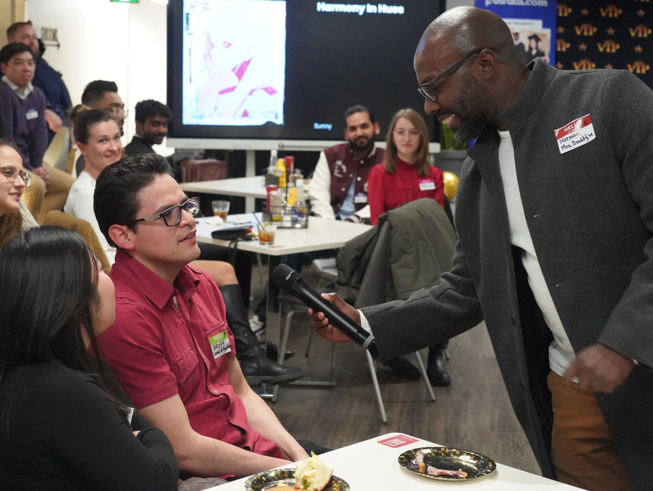A man with glasses and a maroon shirt is speaking into a microphone held by a man with a beard, glasses, and a gray blazer at a social gathering. Several people are seated around a table, smiling and listening. A large screen in the background displays an illustration of a woman, and there are various drinks, snacks, and decorations on the tables.