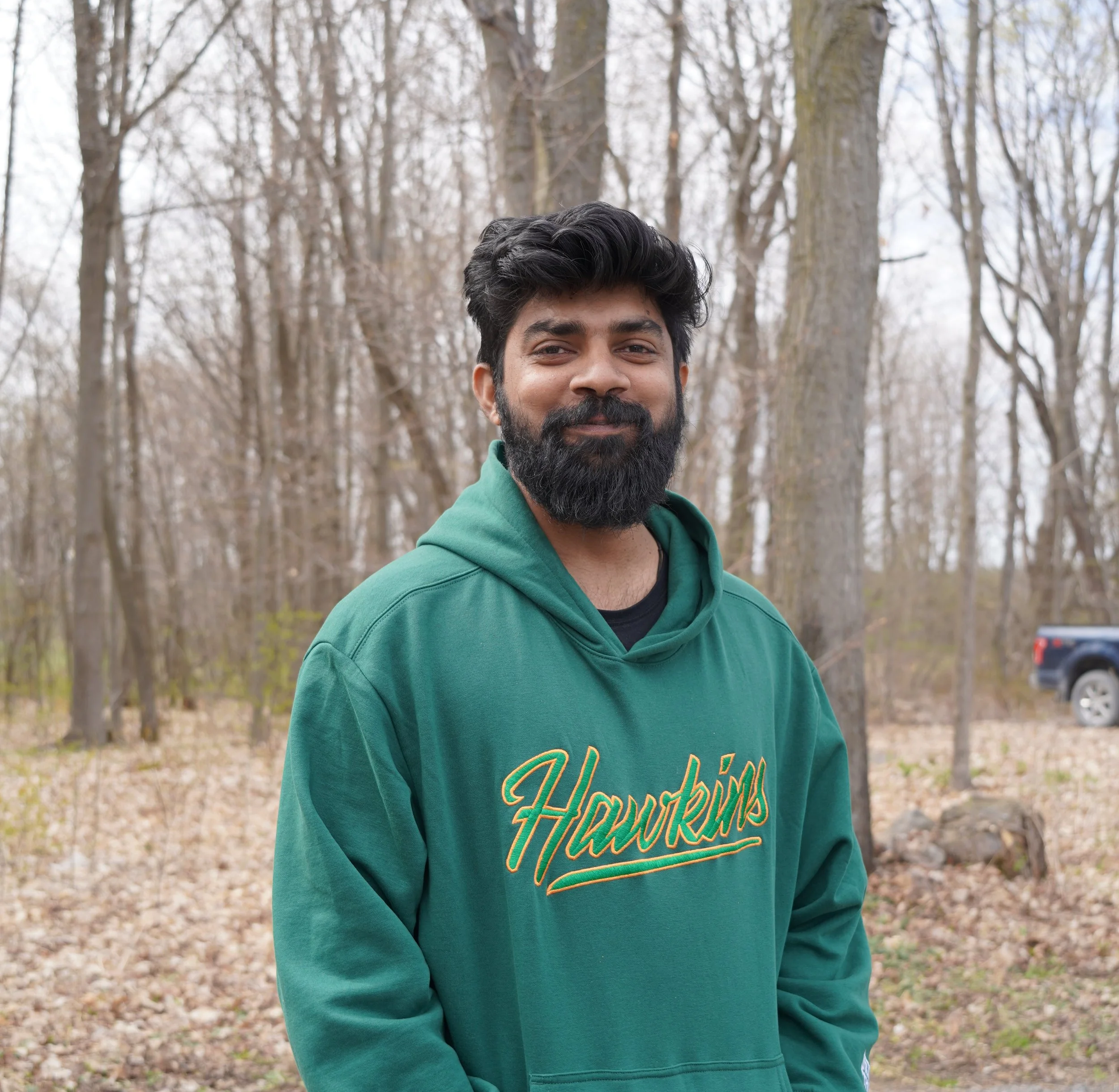 A man with dark hair and a beard smiles in an outdoor wooded area during late fall or early spring.
