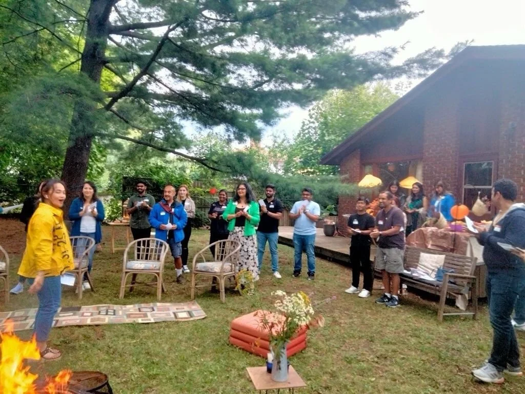 Group of people gathered outdoors around a small fire for a celebration, with some holding drinks, in a backyard with trees and a brick house.