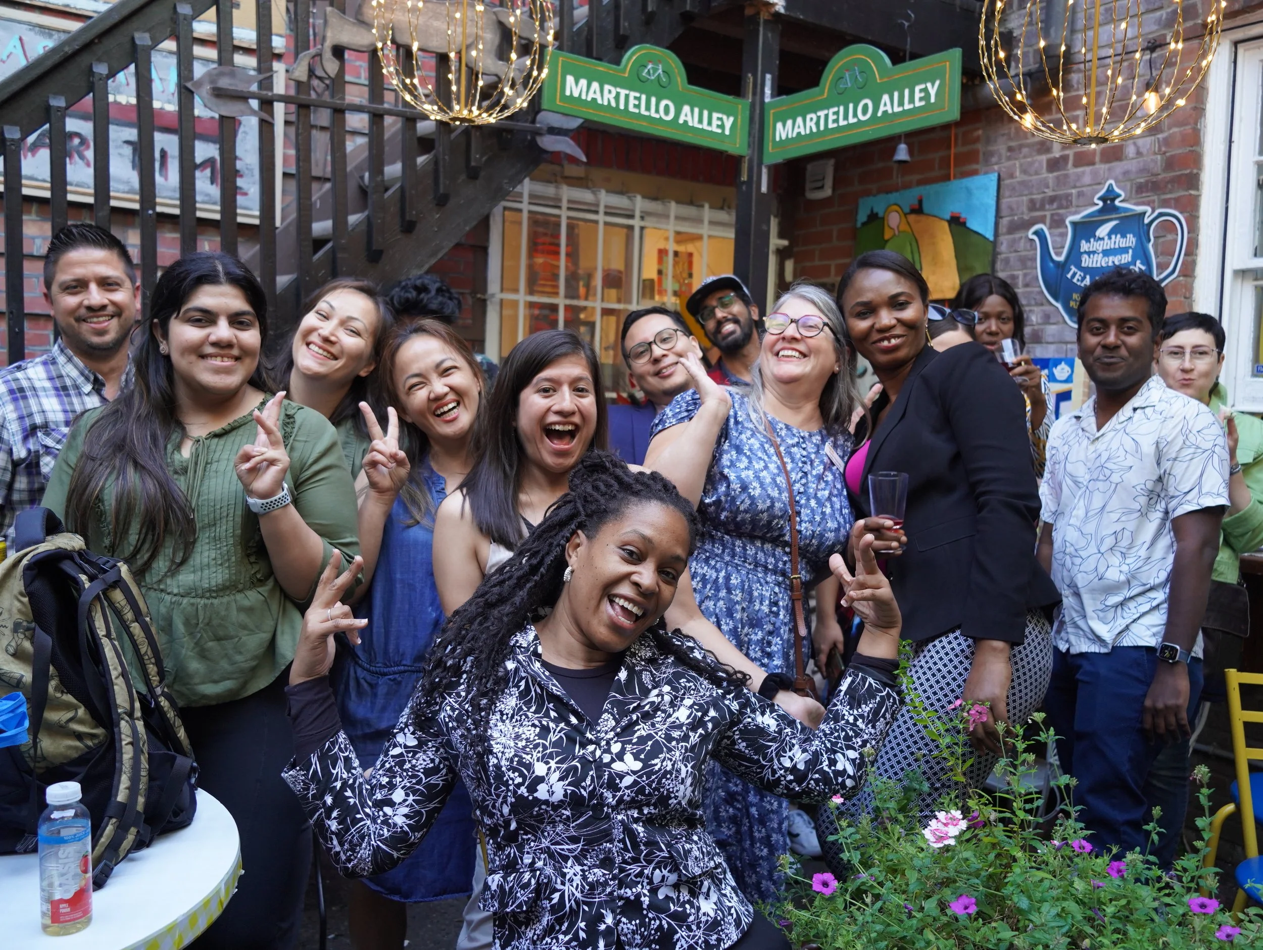 A group of diverse people enjoying a gathering outside, some smiling and making peace signs, with a brick building, string lights, and street signs in the background.
