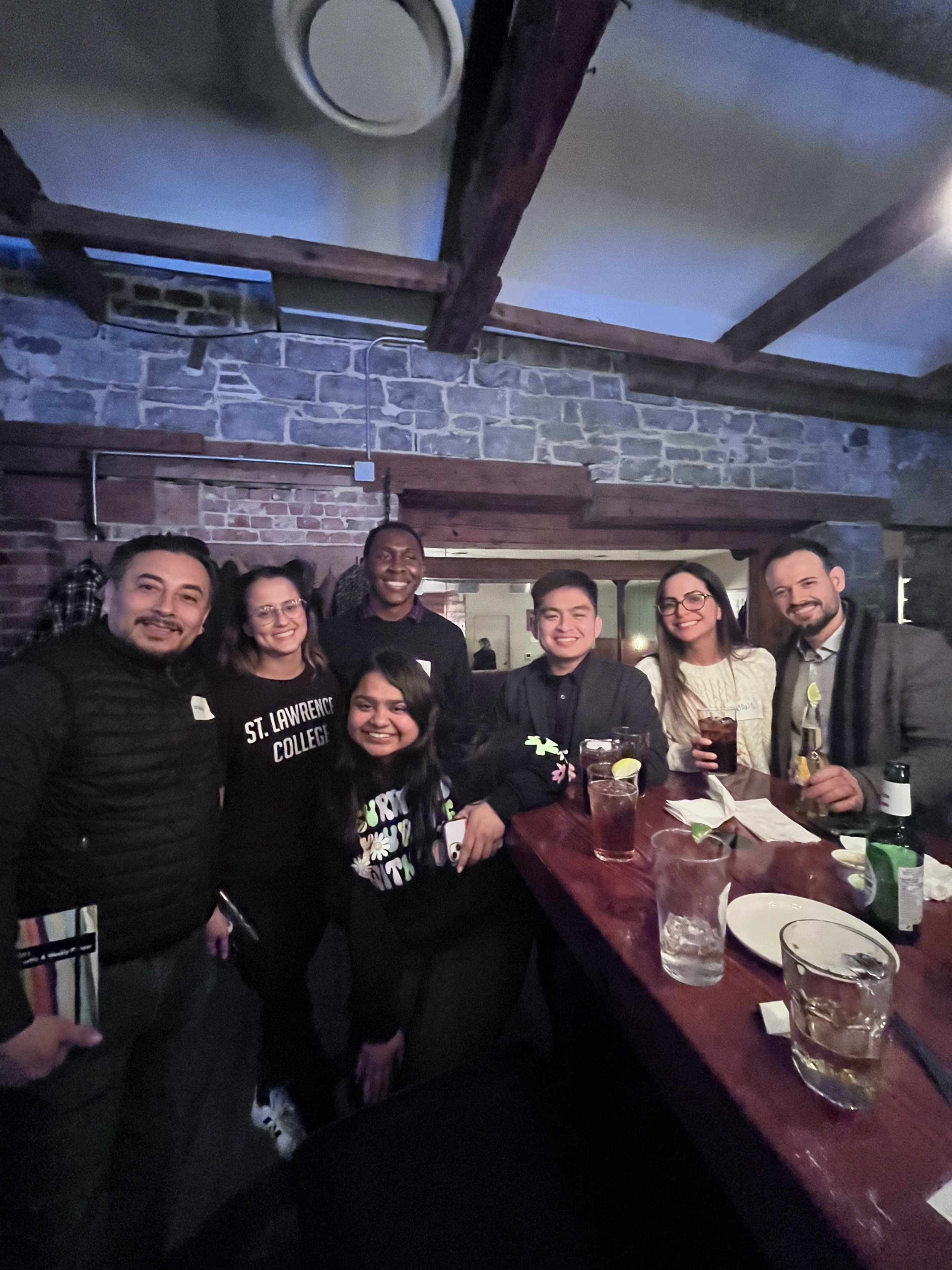 Group of seven friends smiling and posing for a photo at a bar or restaurant, with drinks on the counter, in a cozy setting with exposed brick and wooden beams.