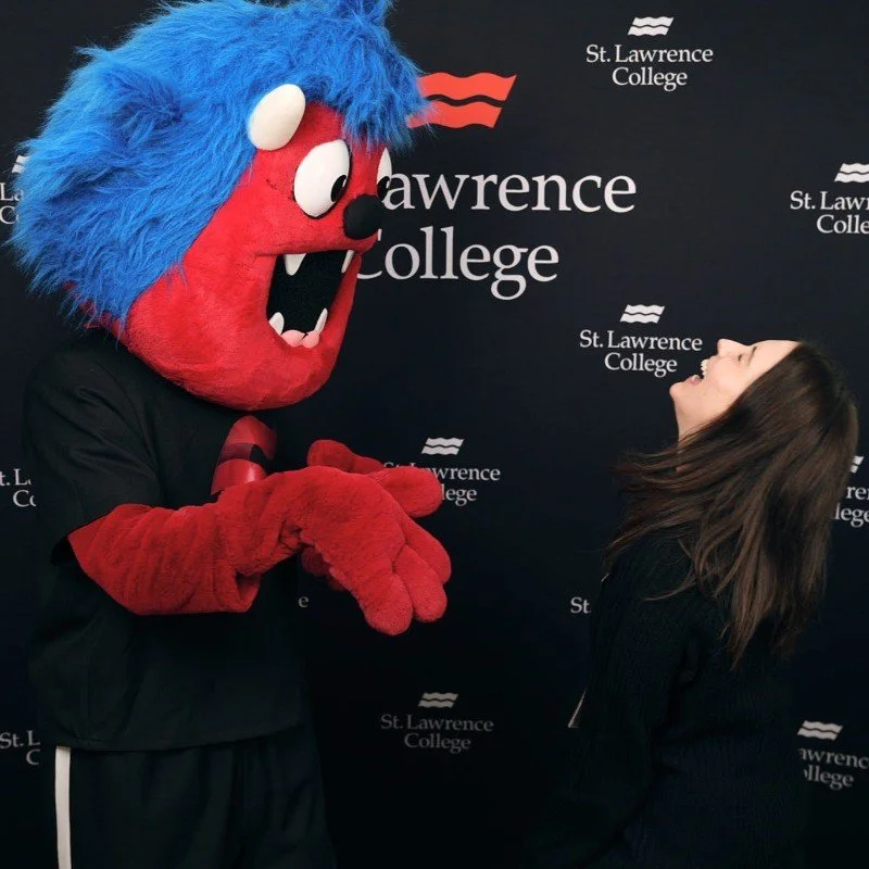 Person in a large red monster mascot costume with blue hair and white eyes talking to a smiling woman at St. Lawrence College event.