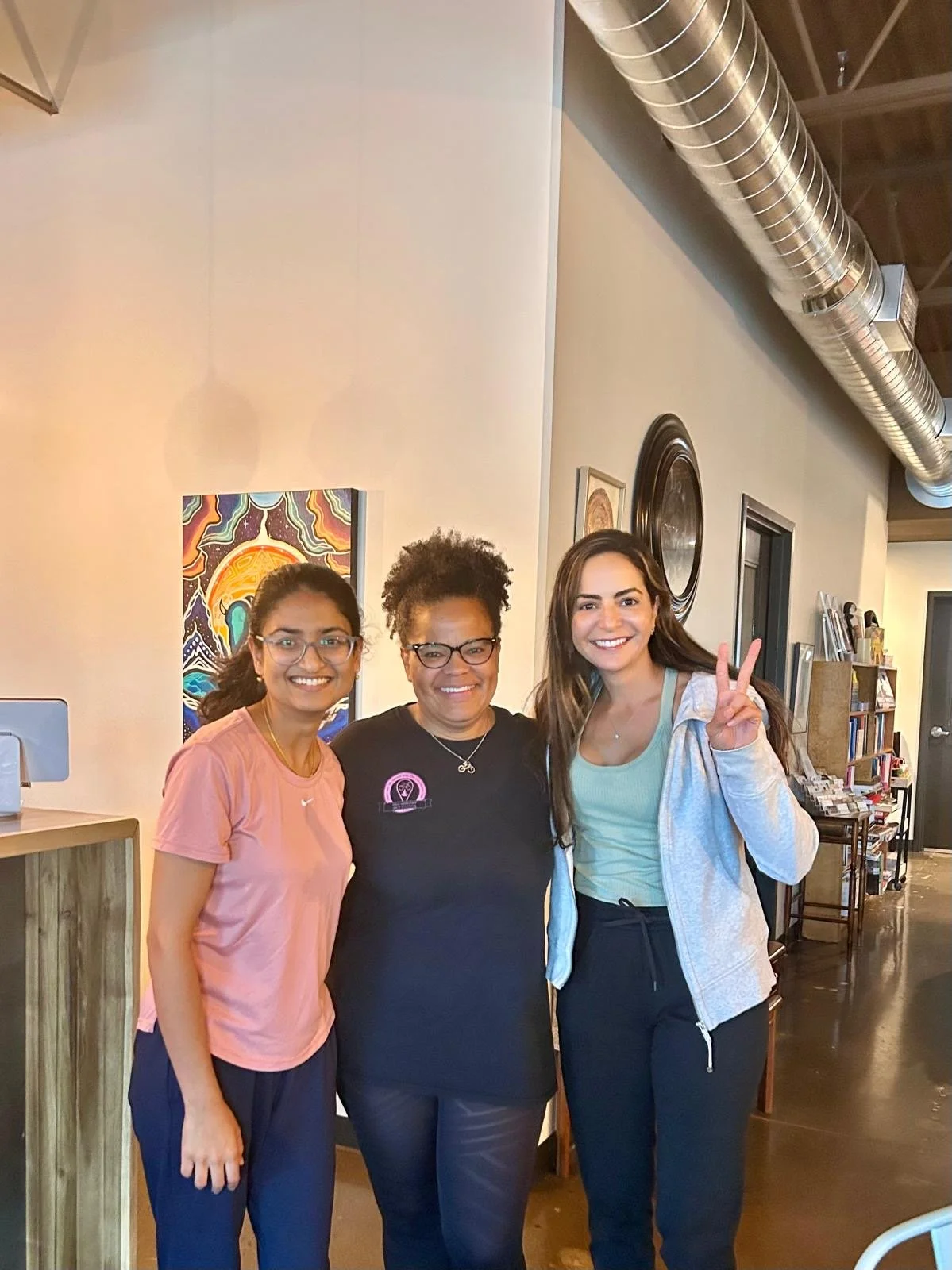 Three women standing close together indoors, smiling at the camera. The woman on the right is making a peace sign with her fingers.