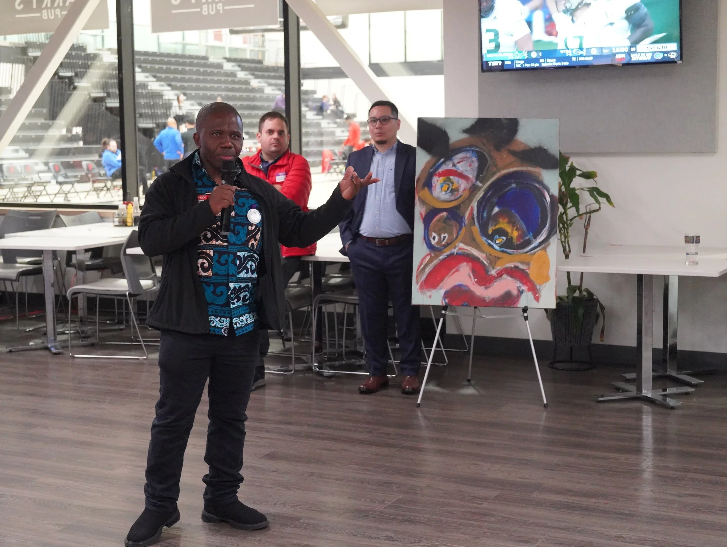 A man holding a microphone speaking in front of an audience with a large abstract painting on an easel and two other men standing behind him, in a spacious room with large windows and a TV screen showing a sports game.