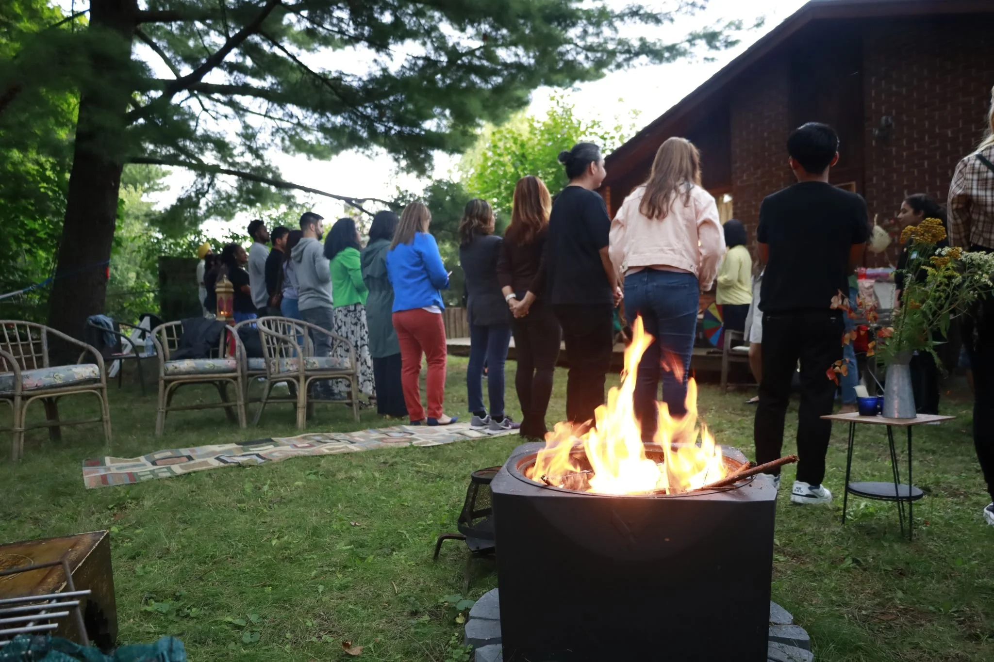 A group of people standing in a line outdoors in a backyard with trees, facing a building. There is a firepit in the foreground and chairs around the yard.