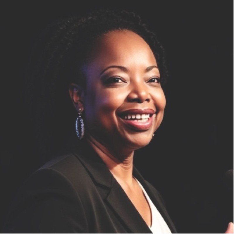 A smiling African American woman with short curly hair, wearing earrings and a black blazer, photographed against a dark background.