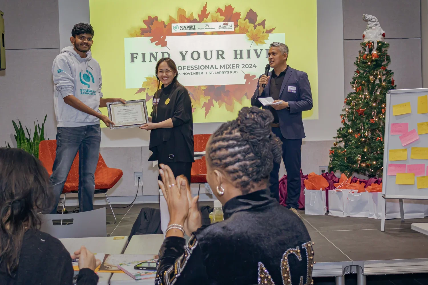 A man in a hoodie receives a framed certificate from a woman in black at an award ceremony. A man in a suit with a microphone stands nearby. The background has a yellow banner with fall leaves and the text 'Find Your Hive Professional Mixer 2024'. There is a decorated Christmas tree and gift bags on the stage, with audience members clapping.