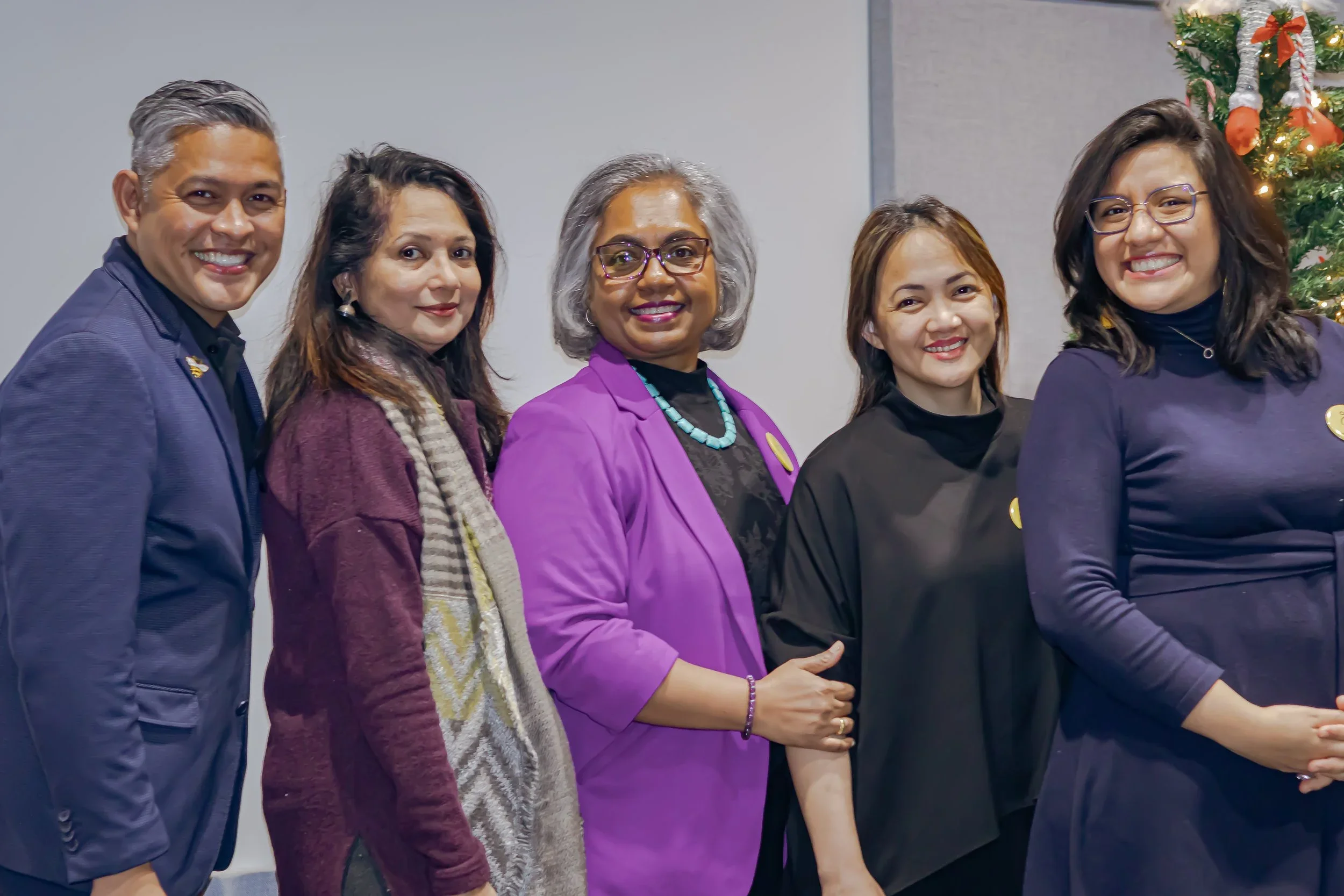 Group of five women standing together at a festive event, with a Christmas tree decorated with ornaments in the background.