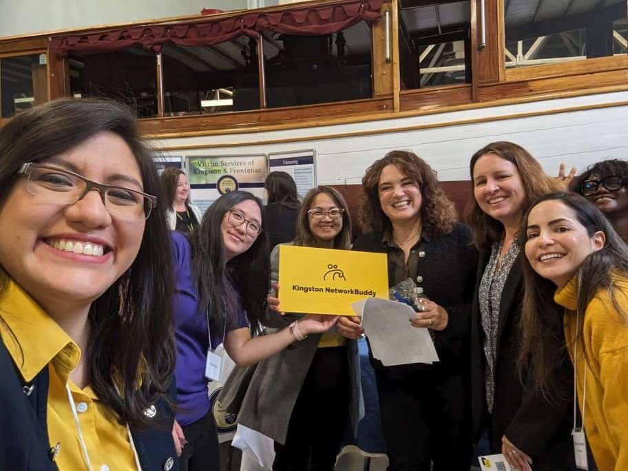 Group of women smiling and holding a yellow sign that reads 'Kingston NetworkBuddy' at an indoor event.