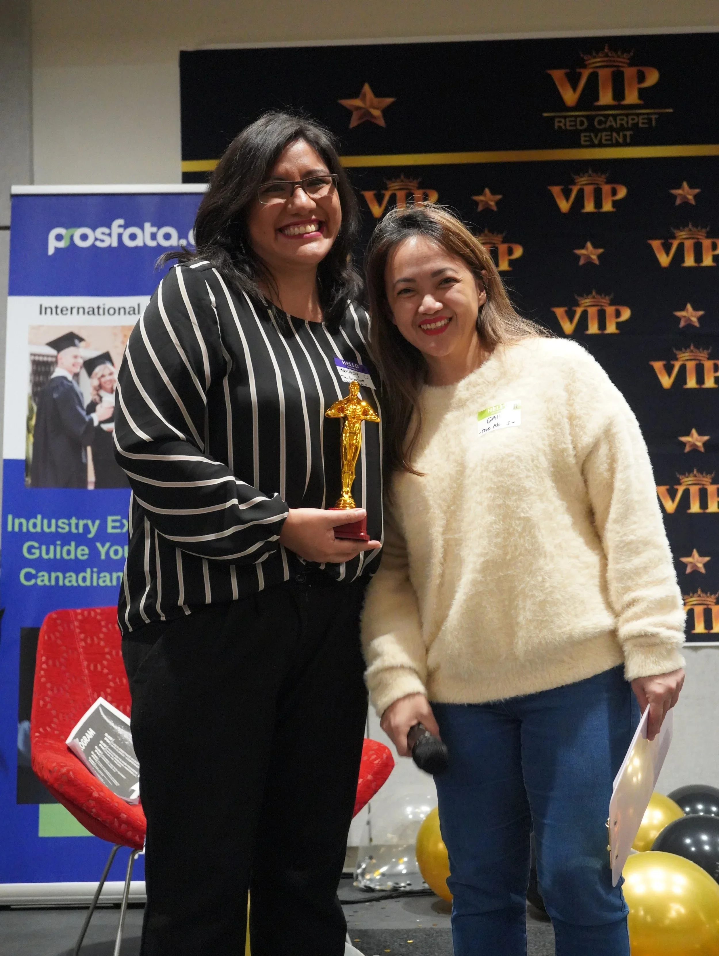 Two women standing on a stage, smiling at the camera. One woman is holding a gold trophy. The backdrop has VIP and red carpet event logos, balloons are on the floor, and a microphone is in the second woman's hand.