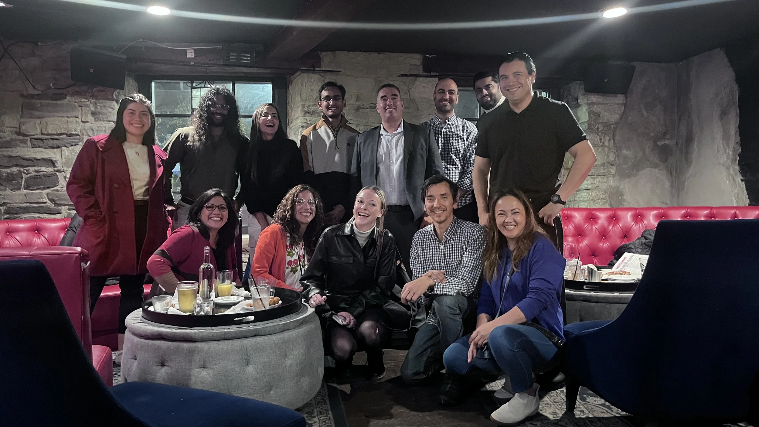 Group of fifteen diverse people smiling and posing for a photo in a cozy restaurant or bar with stone walls and modern furniture.