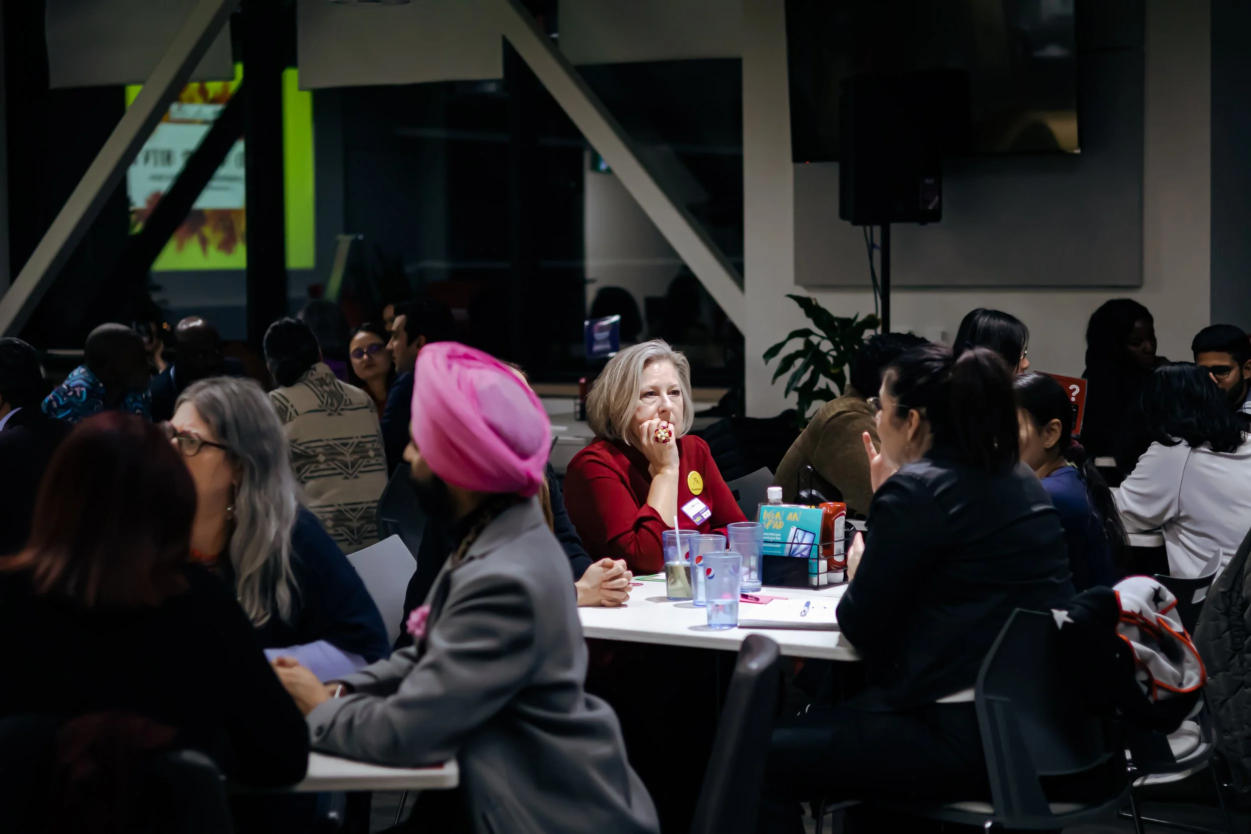 Group of diverse people attending a conference or meeting in a large room, sitting at tables, some engaged in conversation, with a woman in a red blazer sitting in the middle, resting her hand on her face, and a woman in a pink headscarf next to her.
