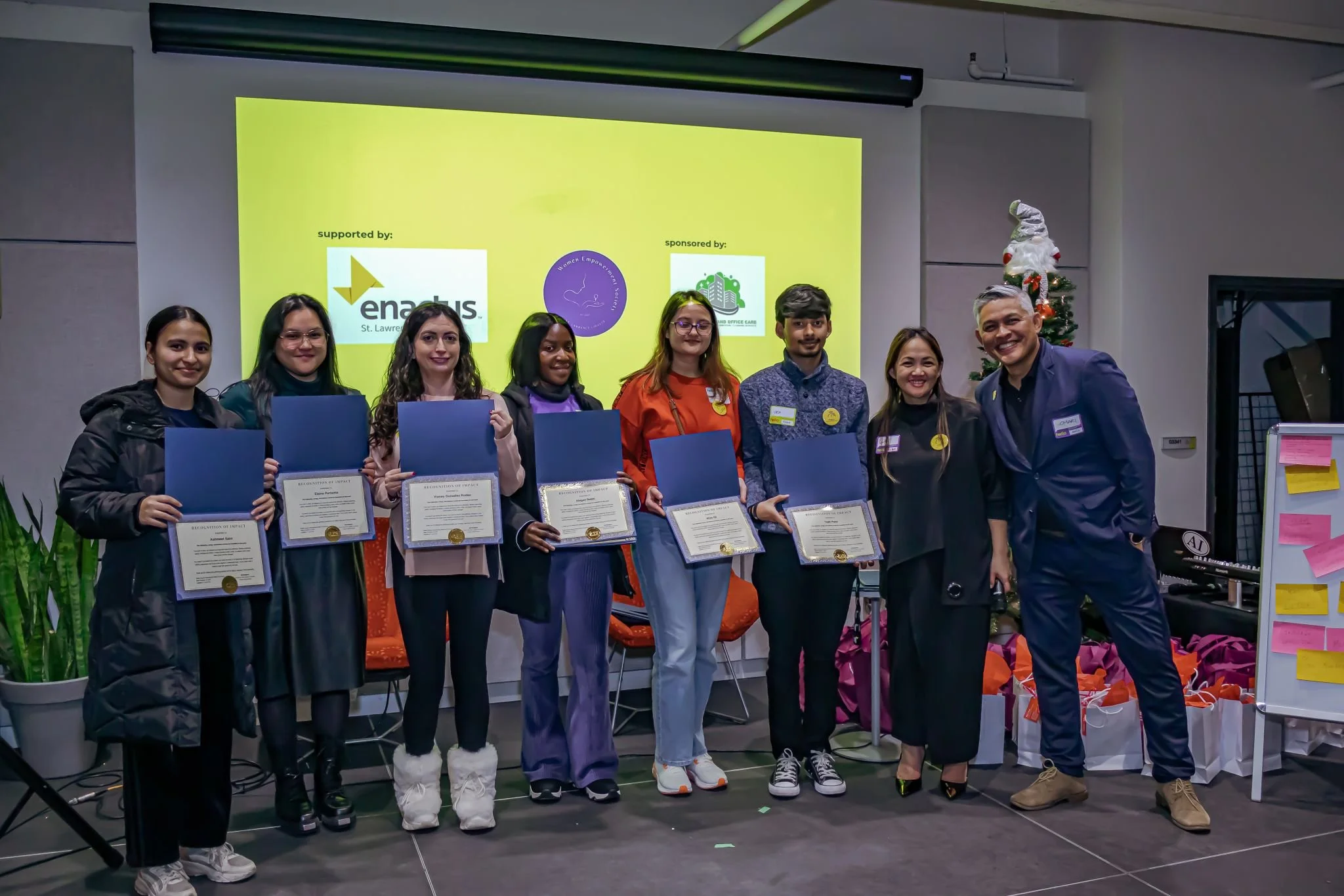 Group of seven women and one man standing indoors, each holding a certificate or award, with a yellow presentation screen in the background displaying logos and text, and a decorated Christmas tree is visible on the right side.