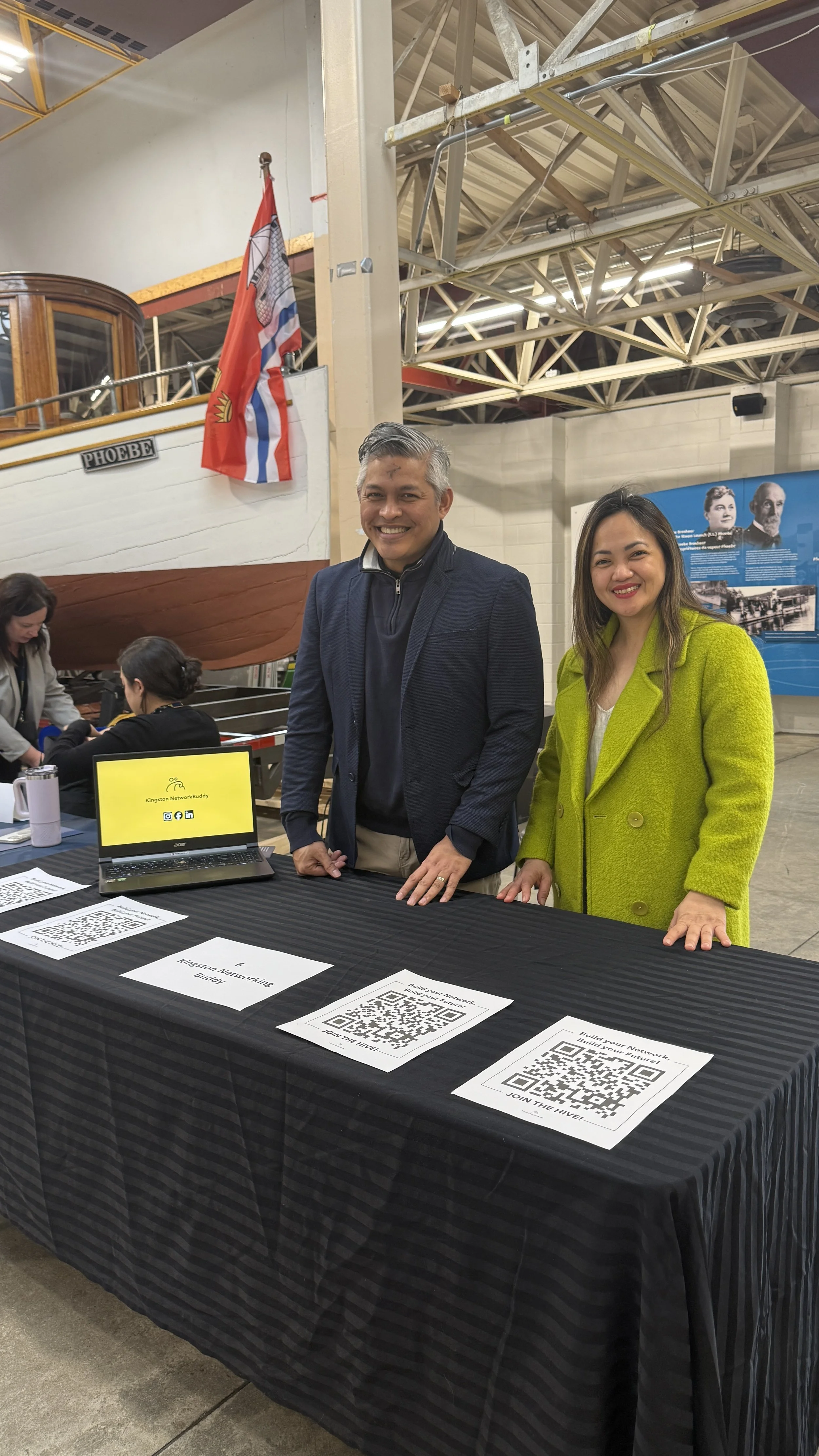 Two smiling people standing behind a table with QR codes at an indoor event. A boat named 'Phoebe' is visible in the background, along with flags and informational displays.