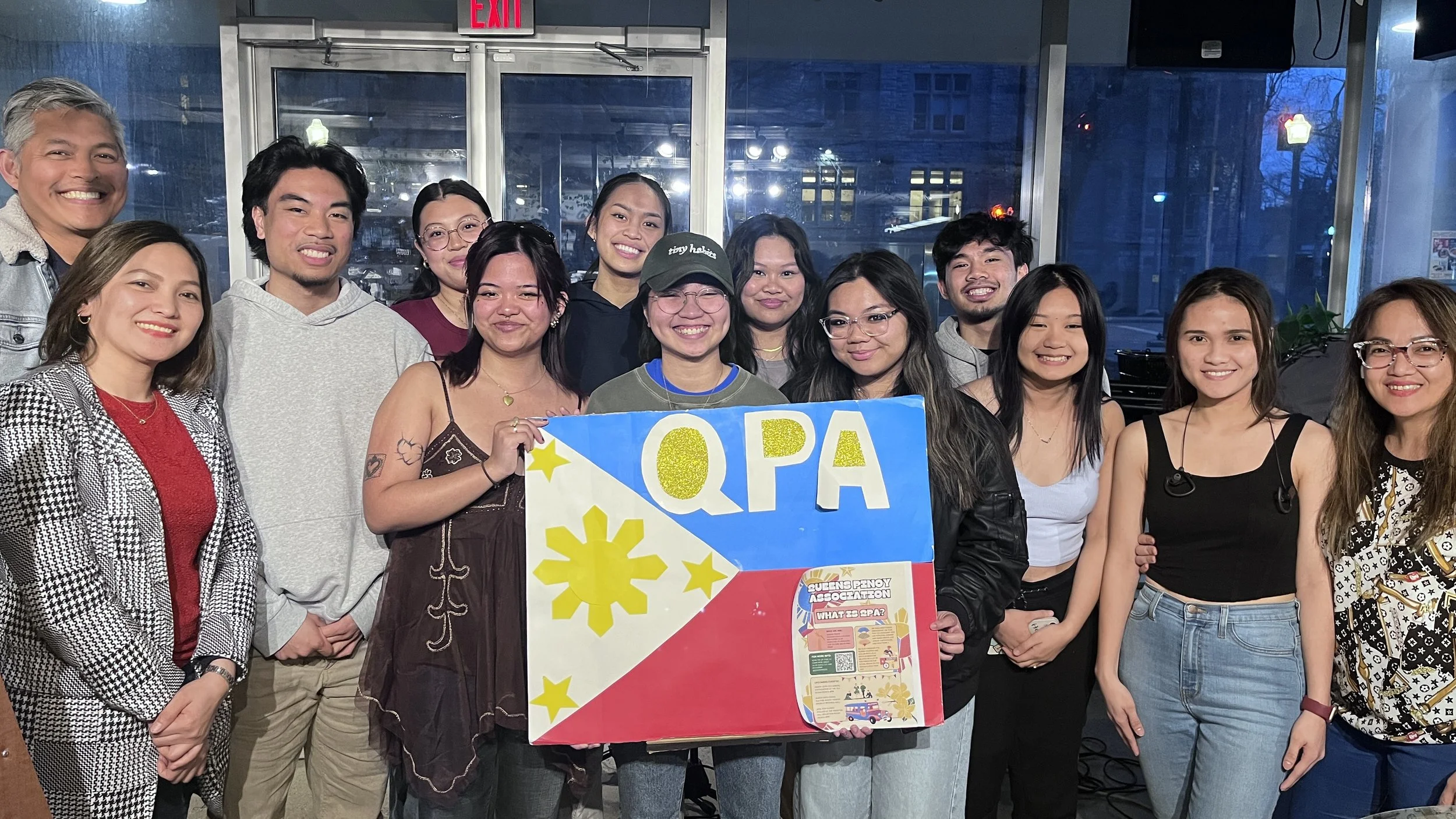 Group of people indoors holding a sign with the letters QPA, surrounded by a diverse crowd smiling for the photo.