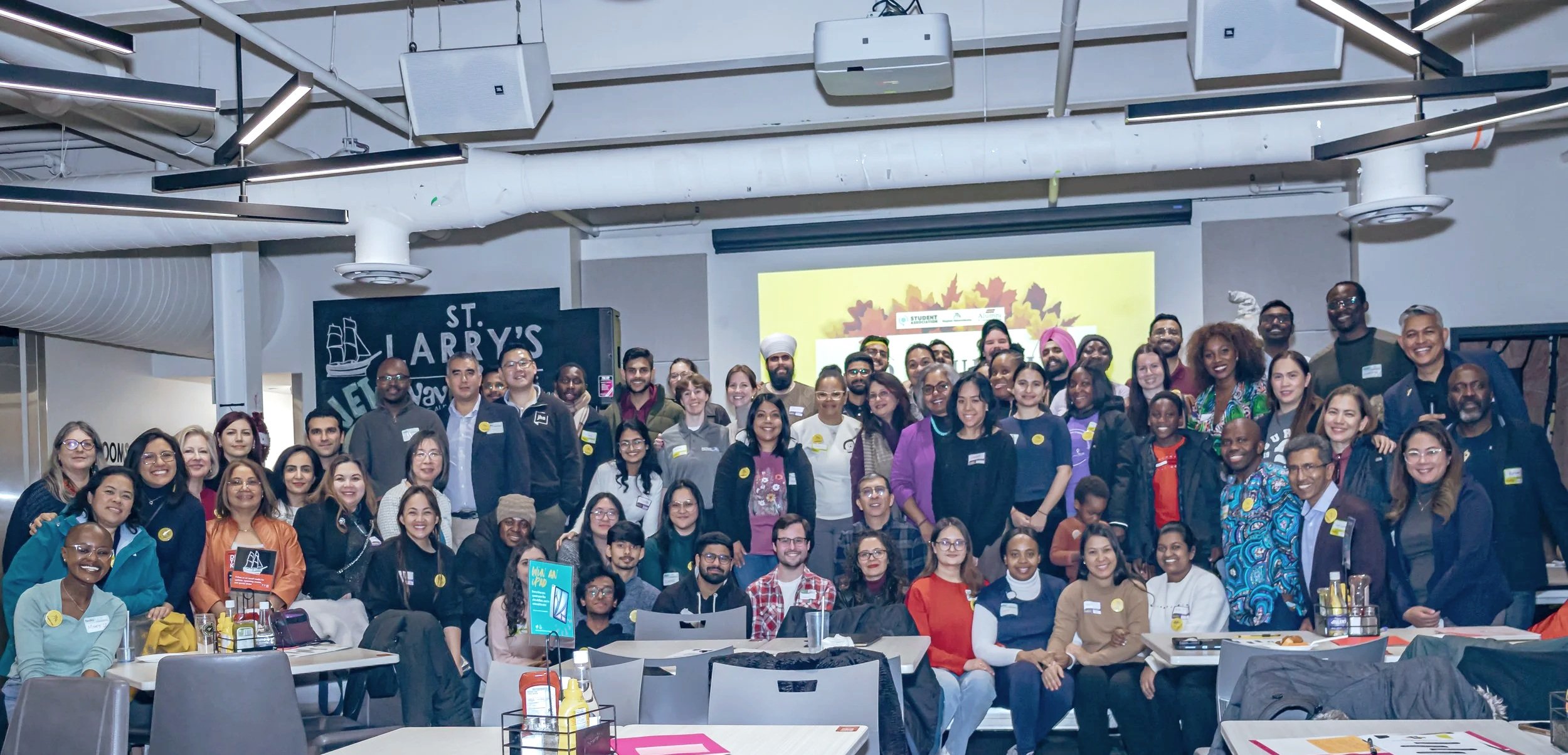 A large group of diverse people gathered in an indoor venue, posing for a group photo. There are tables with drinks and snacks in front, and a projector screen and black sign with white letters saying 'St. Larry's' in the background.