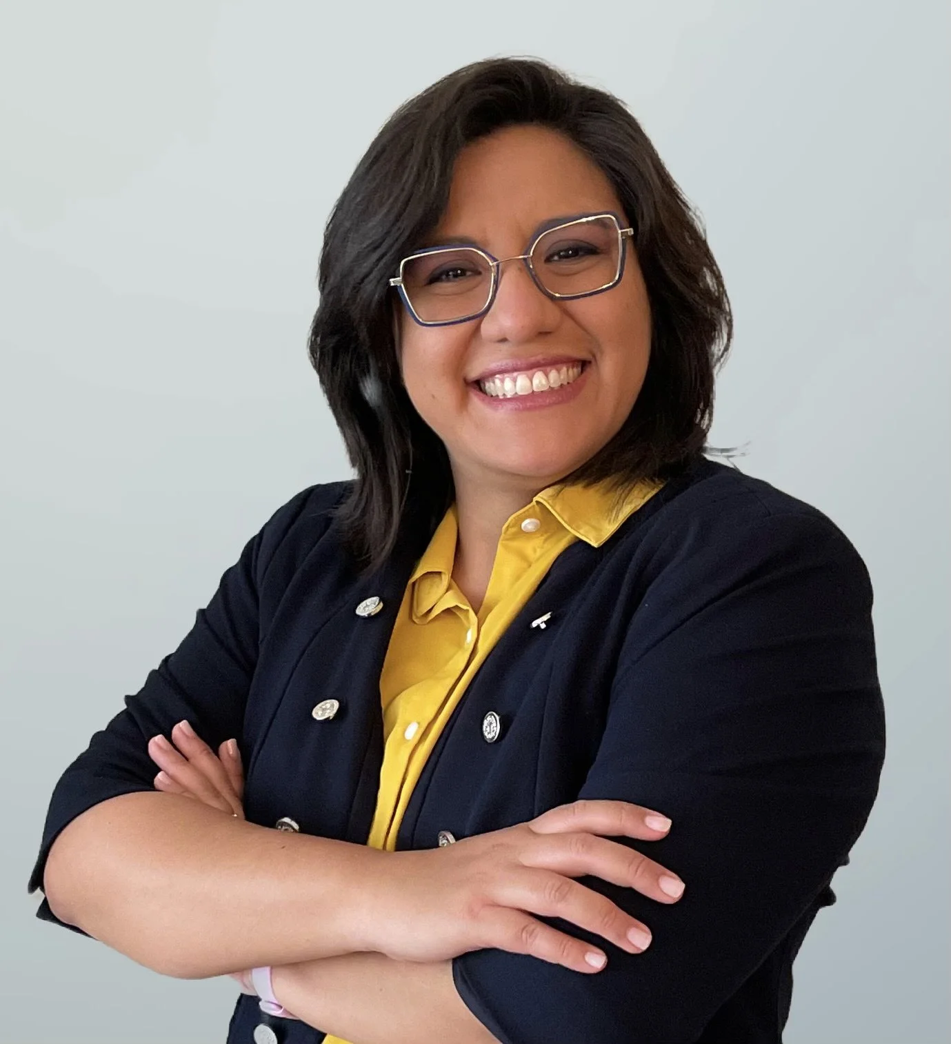 A woman with shoulder-length dark hair and glasses smiling, wearing a yellow blouse and a navy blazer with pins, arms crossed, against a plain light background.