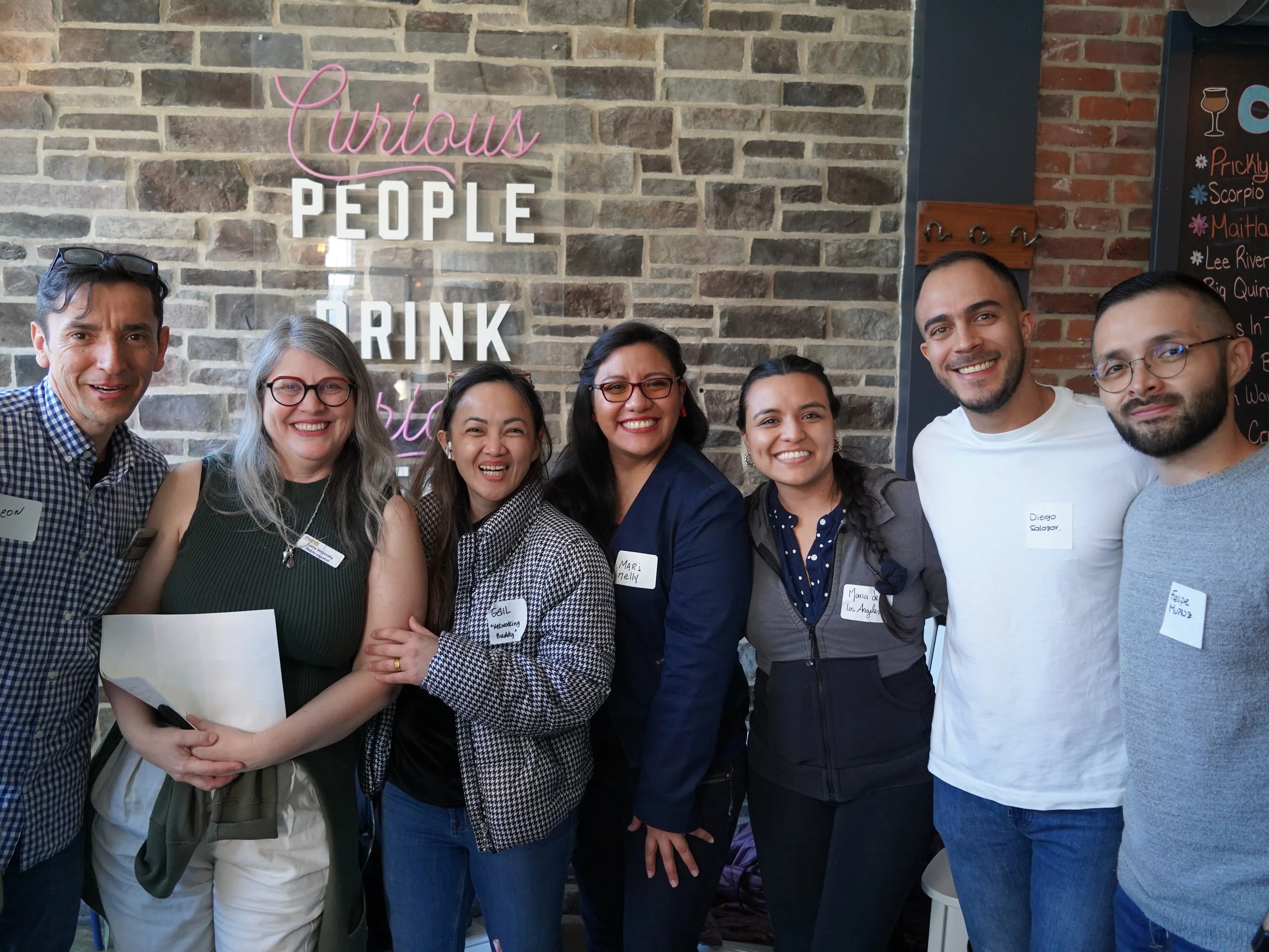 A group of seven diverse smiling people standing together in front of a brick wall with neon sign that reads 'Curious People Drink'.