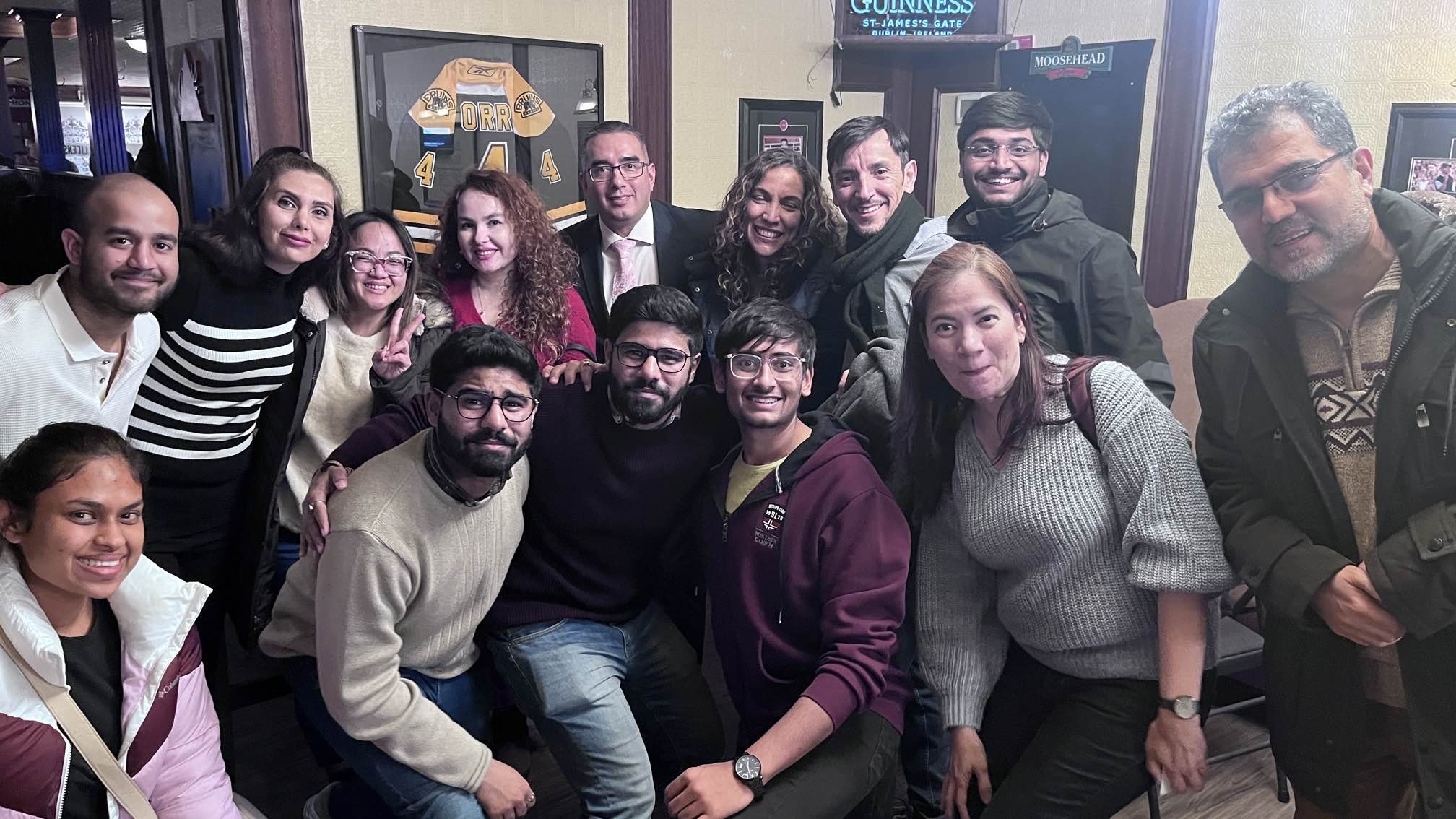 A group of 15 diverse people posing for a photo indoors, smiling and making peace signs, with a framed hockey jersey and pub decor in the background.