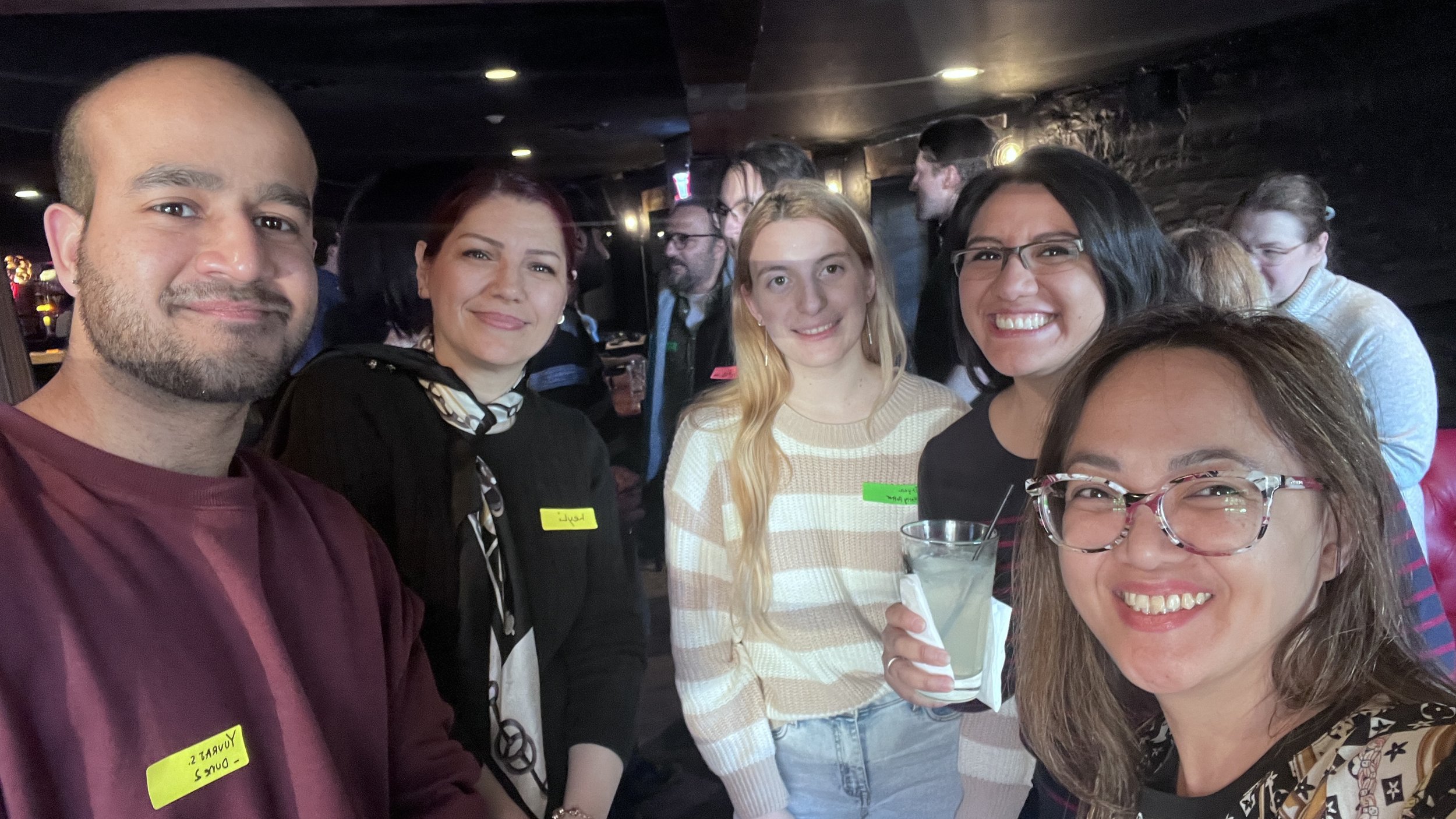 Group of six smiling people at a social gathering in an indoor venue, some holding drinks, with a dark background and warm lighting.