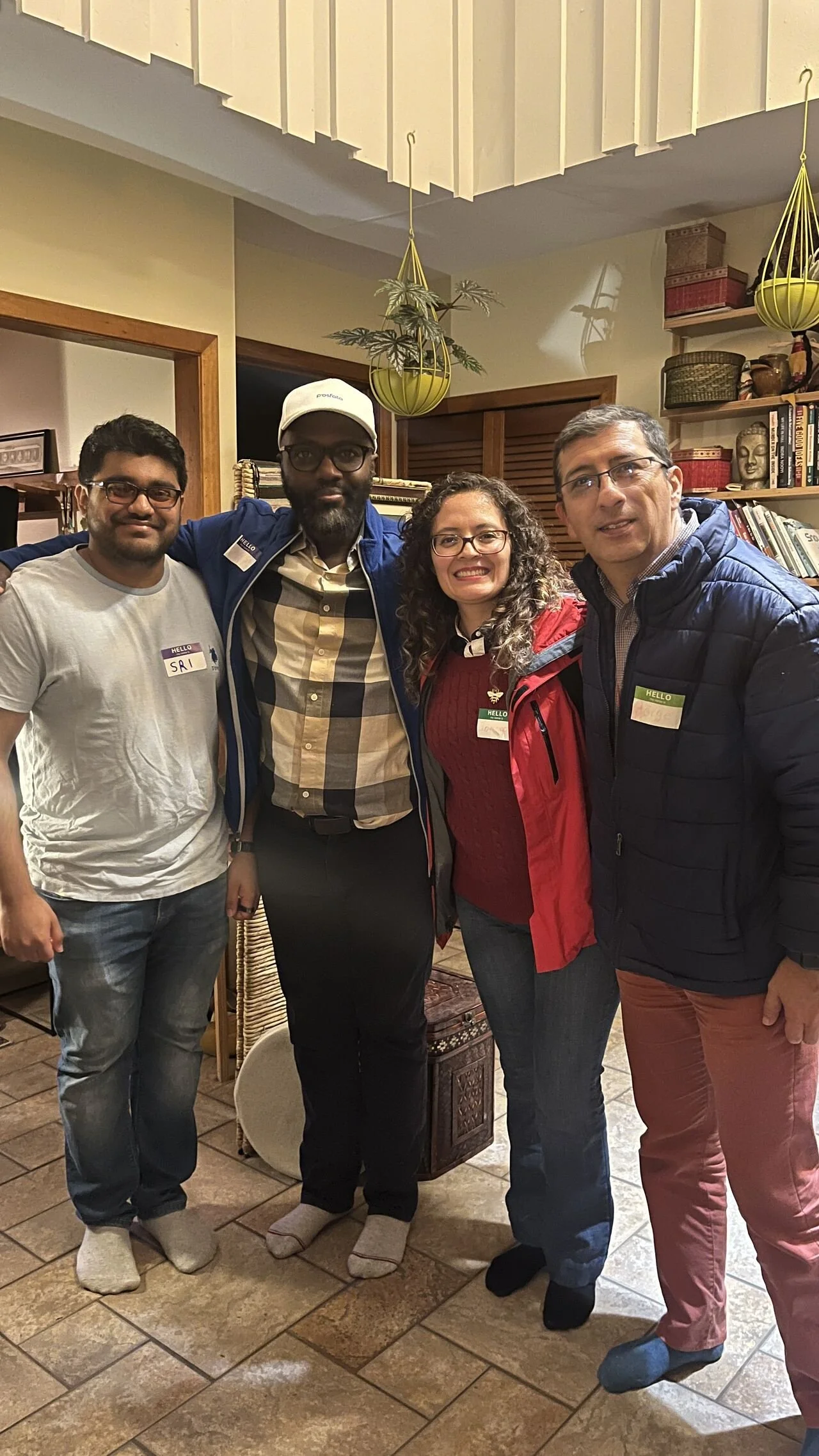Group of four people standing indoors, smiling for the photo, wearing casual clothes and name tags. The background includes a bookshelf, decorative hanging plants, and a wooden door.