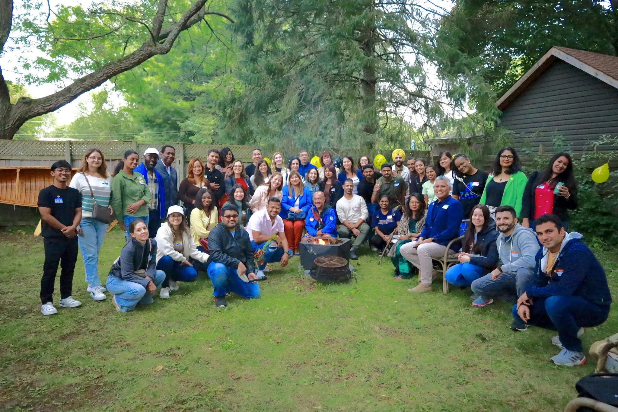 A large group of diverse people gathered outdoors in a backyard with trees, a wooden fence, and a small building, posing for a group photo around a fire pit with balloons and decorations.