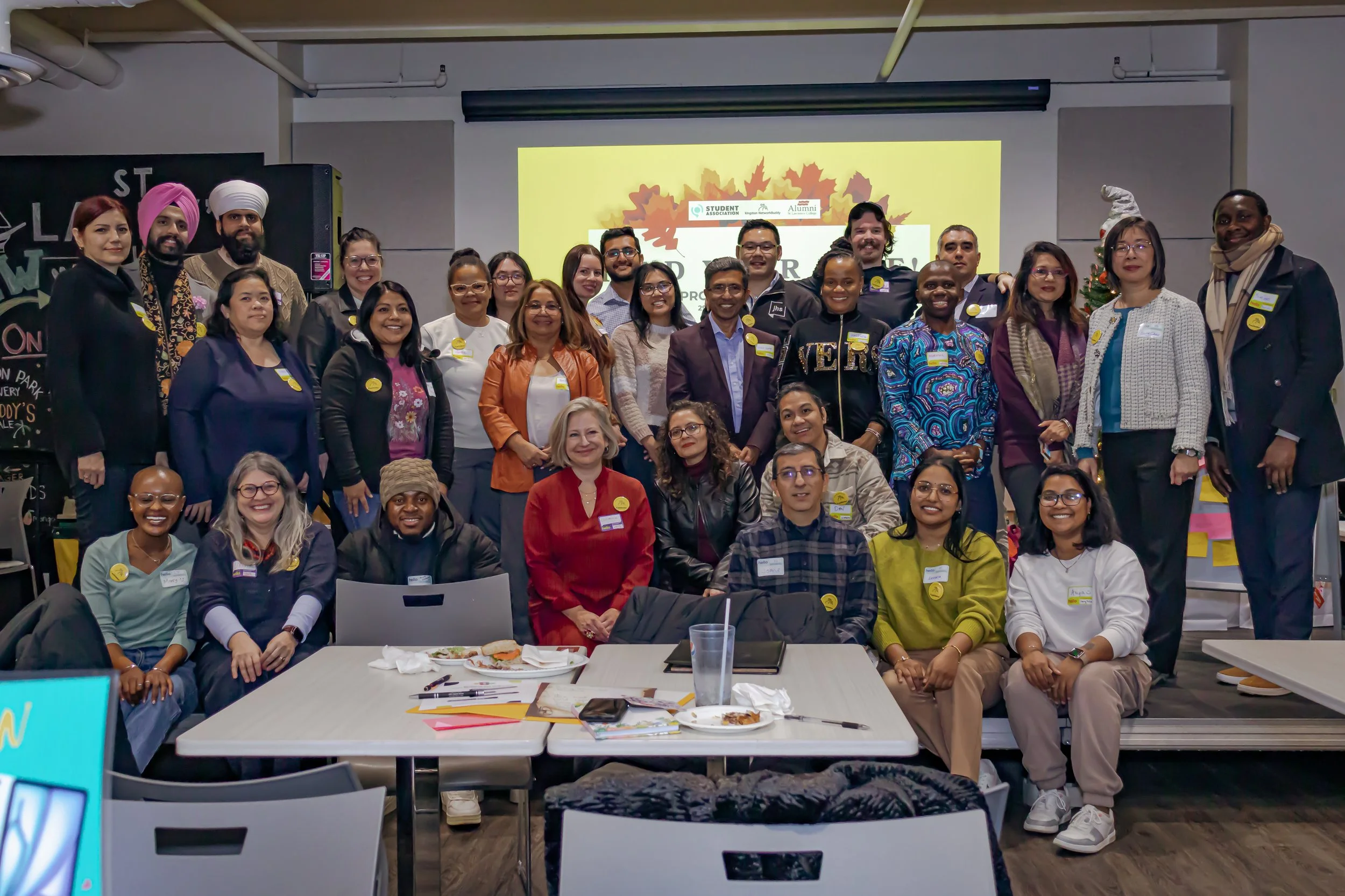 Group of diverse people gathered in a conference room for a presentation or workshop, with some seated at tables and others standing behind, smiling at the camera, and a projector screen displaying autumn leaves in the background.