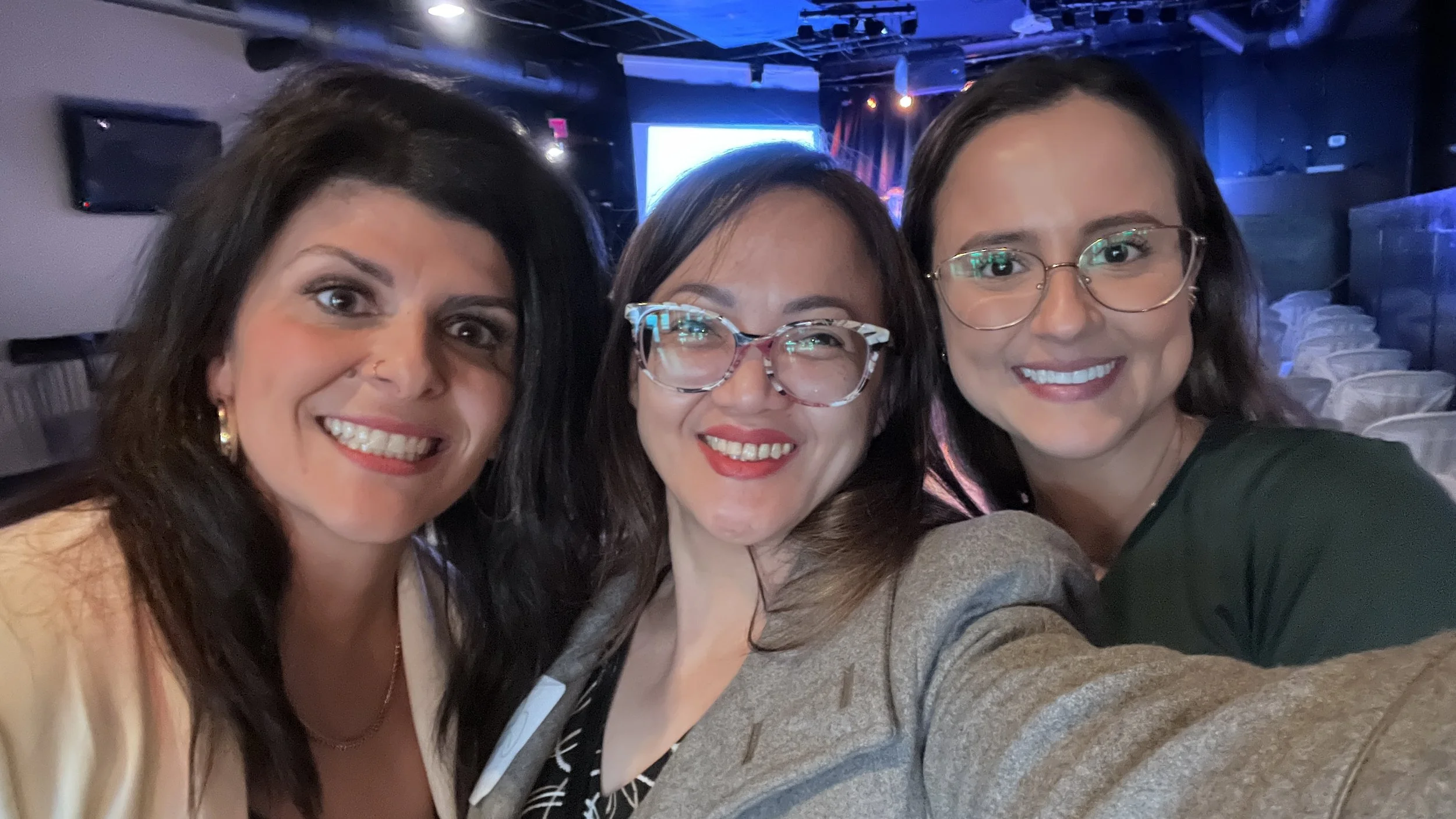 Three women smiling and taking a selfie together indoors at an event or gathering.