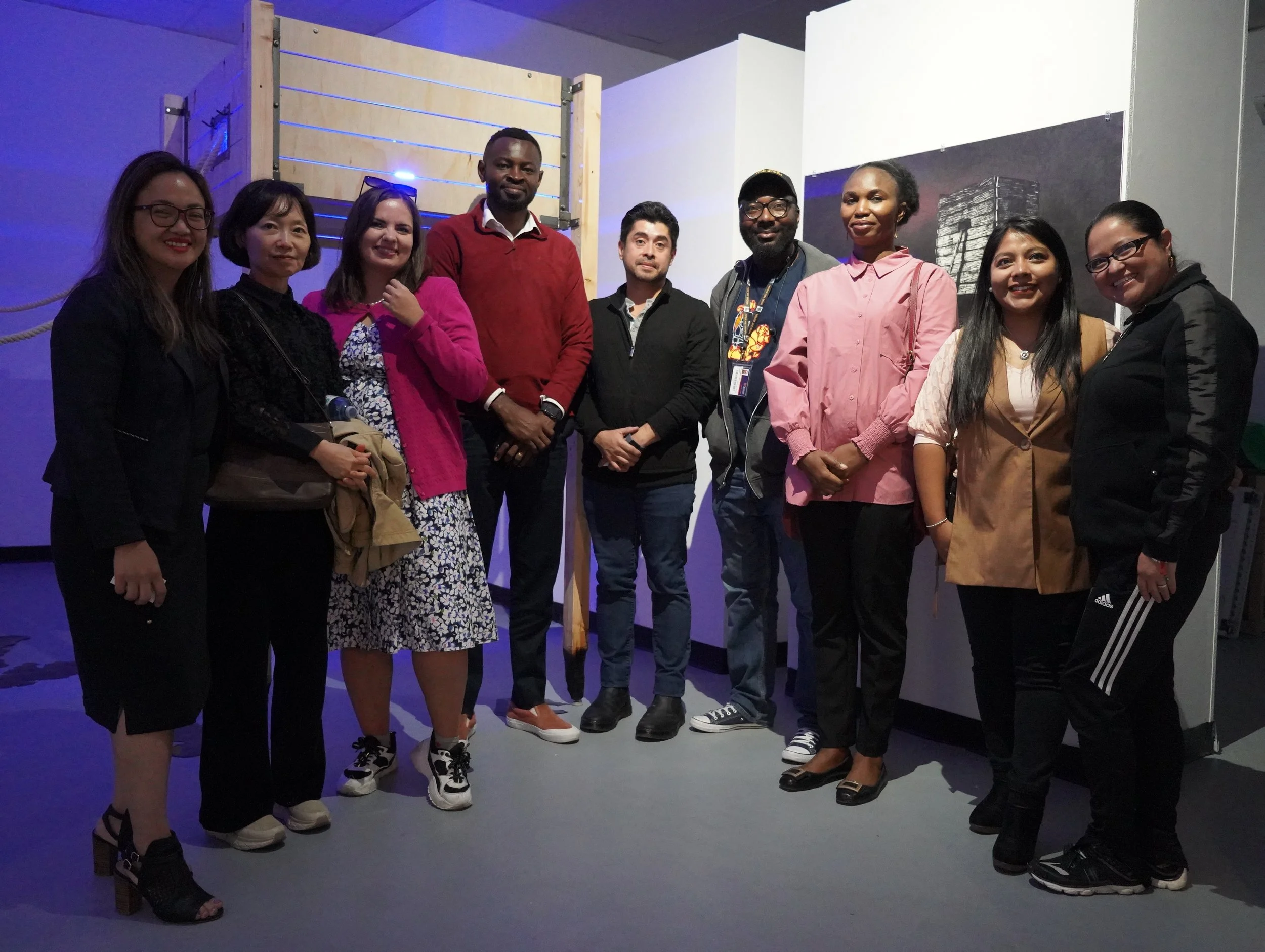 A diverse group of ten people standing together indoors, smiling at the camera, with some holding bags, dressed in casual and semi-formal attire, against a background with a wooden structure and artwork.