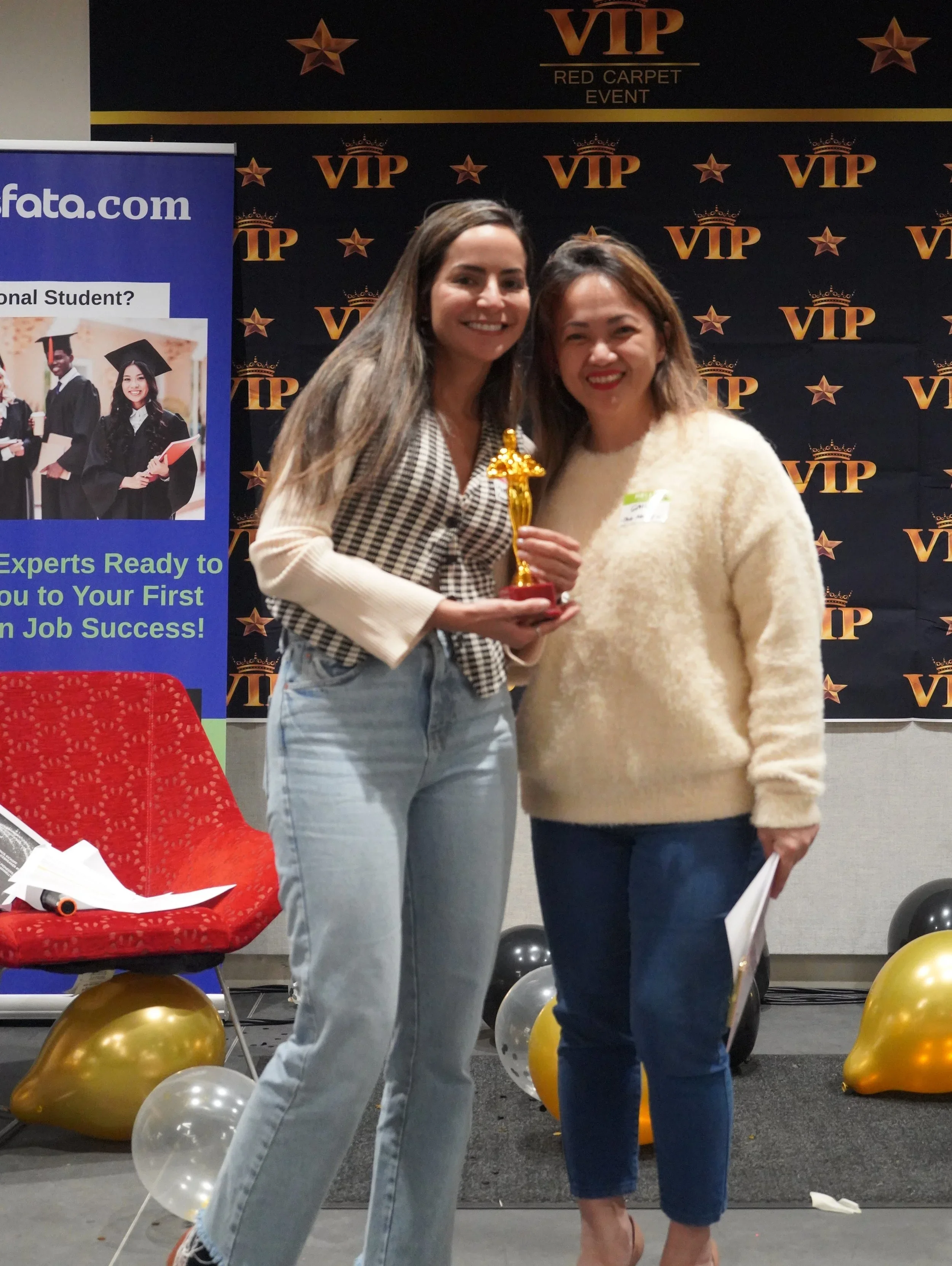 Two women smiling and holding a trophy together at an event, with a VIP backdrop and balloons in the background.