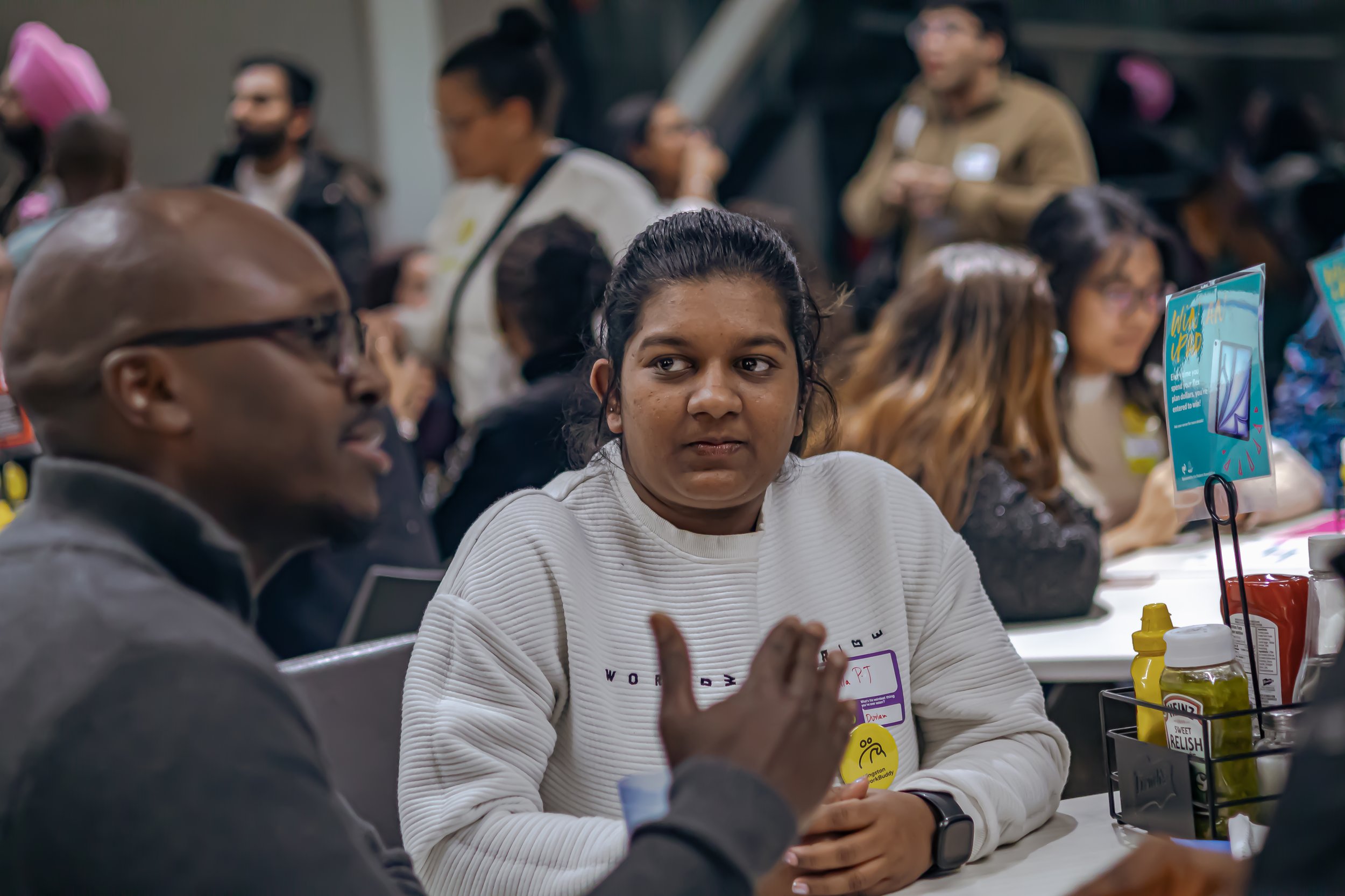 People sitting at a table in a crowded indoor setting, engaged in conversation, with condiments and promotional signs on the table