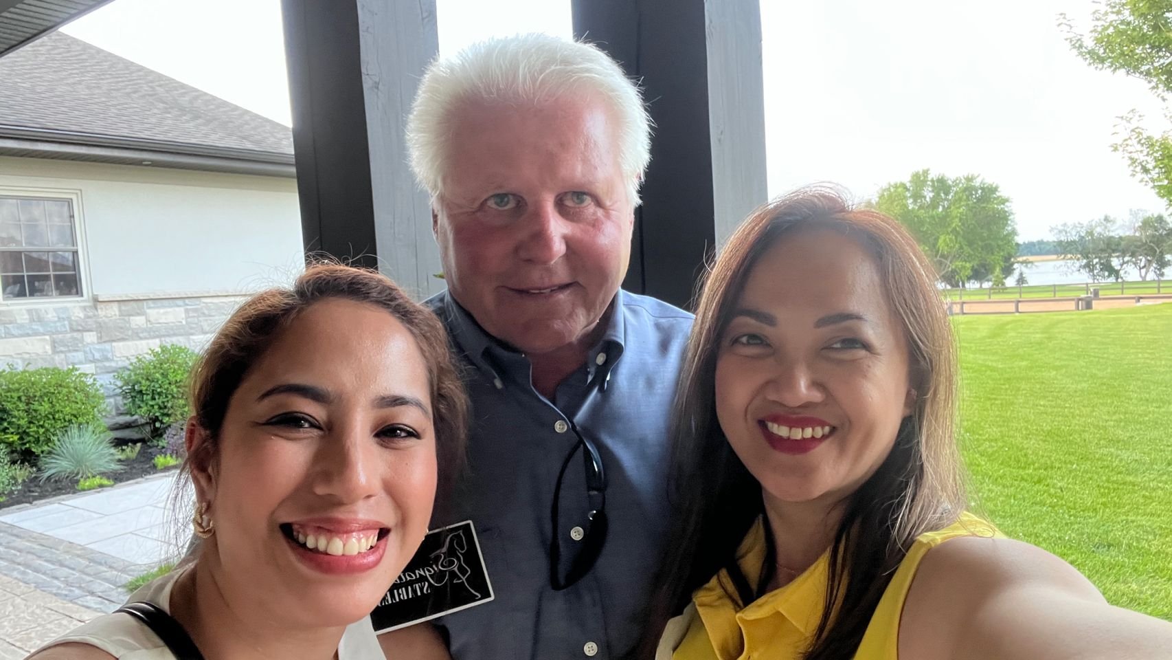 A group of three people smiling for a selfie outdoors with a green lawn, trees, and a body of water visible in the background.