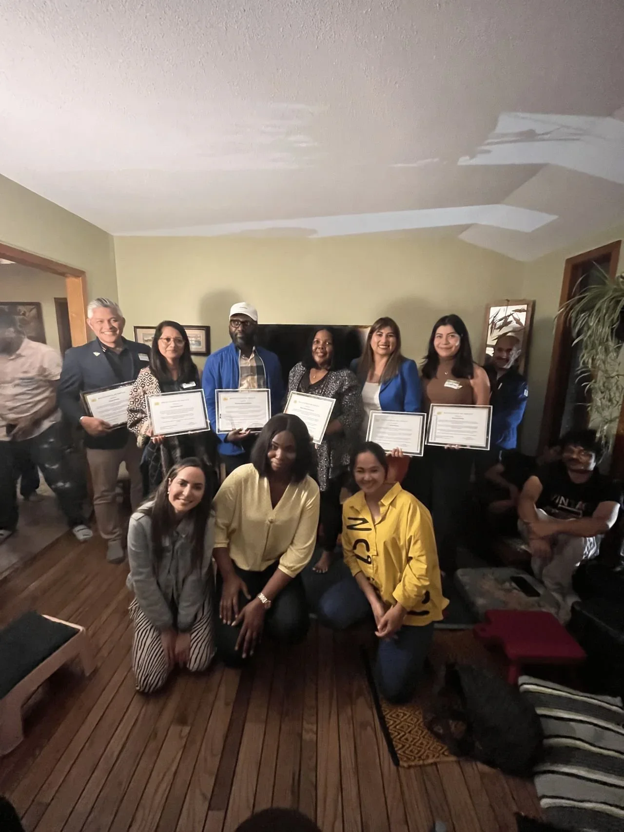 Group of people posing with certificates in a living room, some kneeling in the front and others standing in the back, smiling.