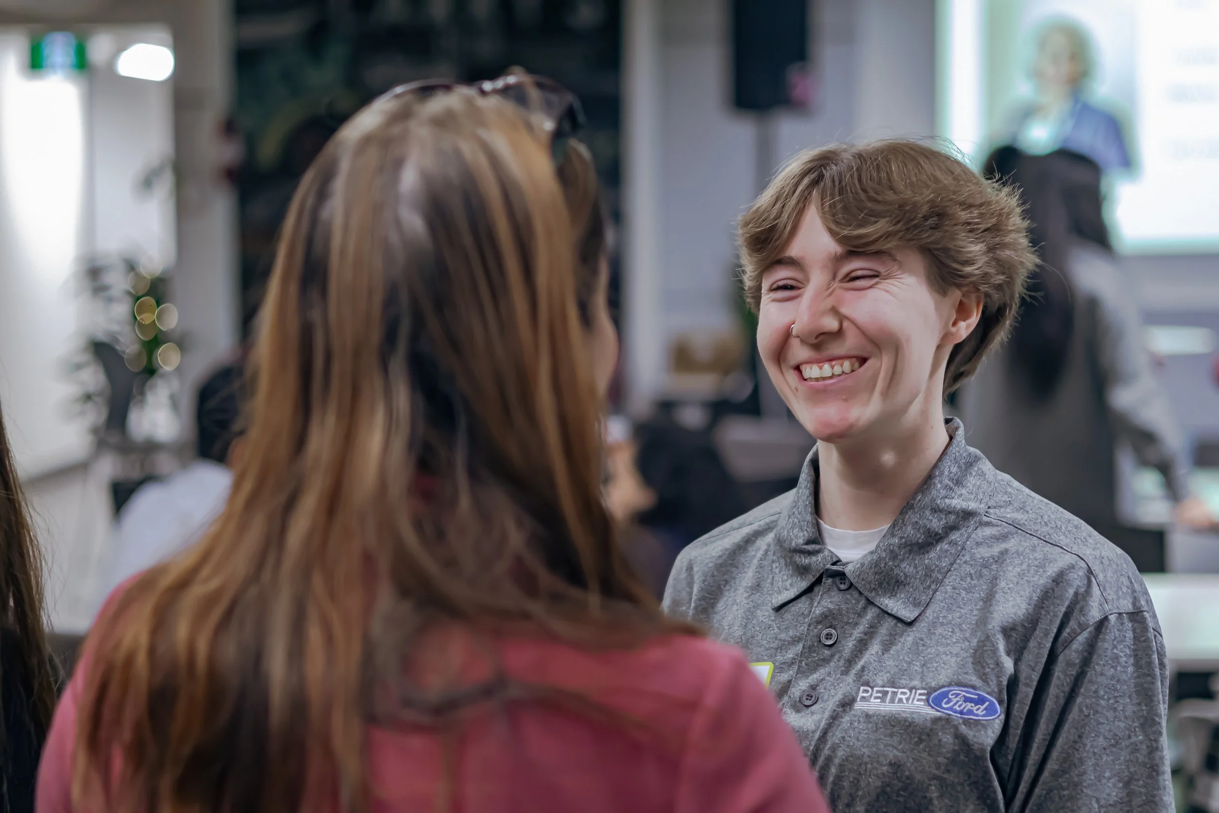 A young man with curly hair smiling and engaging in conversation with a woman at a dealership, with a woman in the background near a presentation screen.