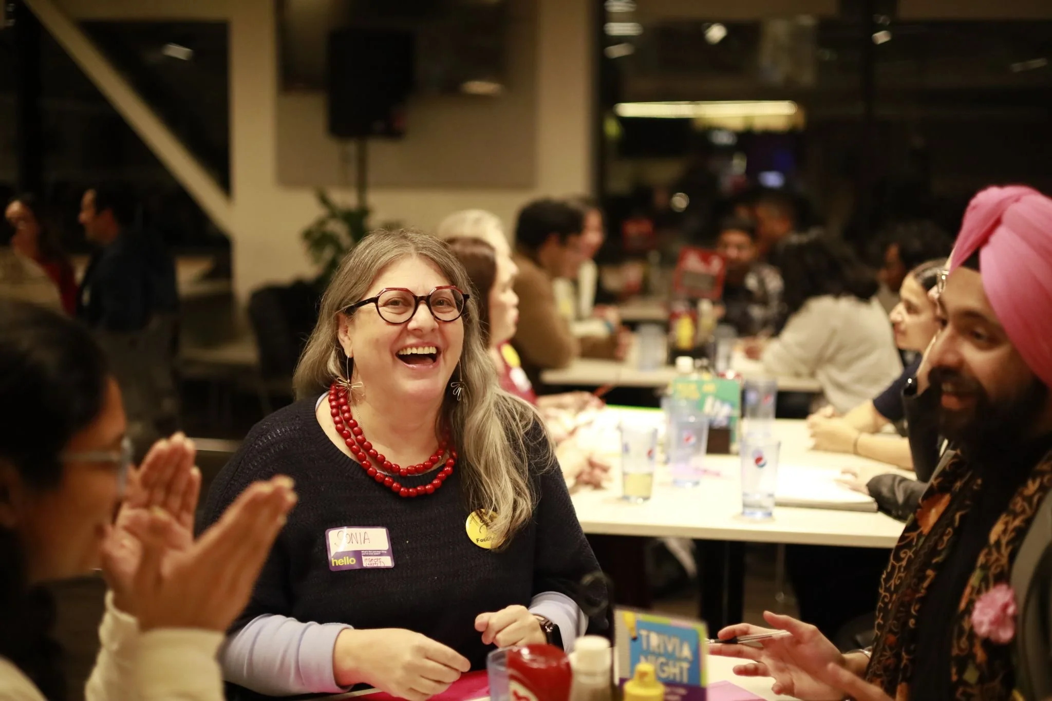 A group of diverse people gathered at tables during a social event, with a smiling woman wearing glasses and a red necklace in the foreground.