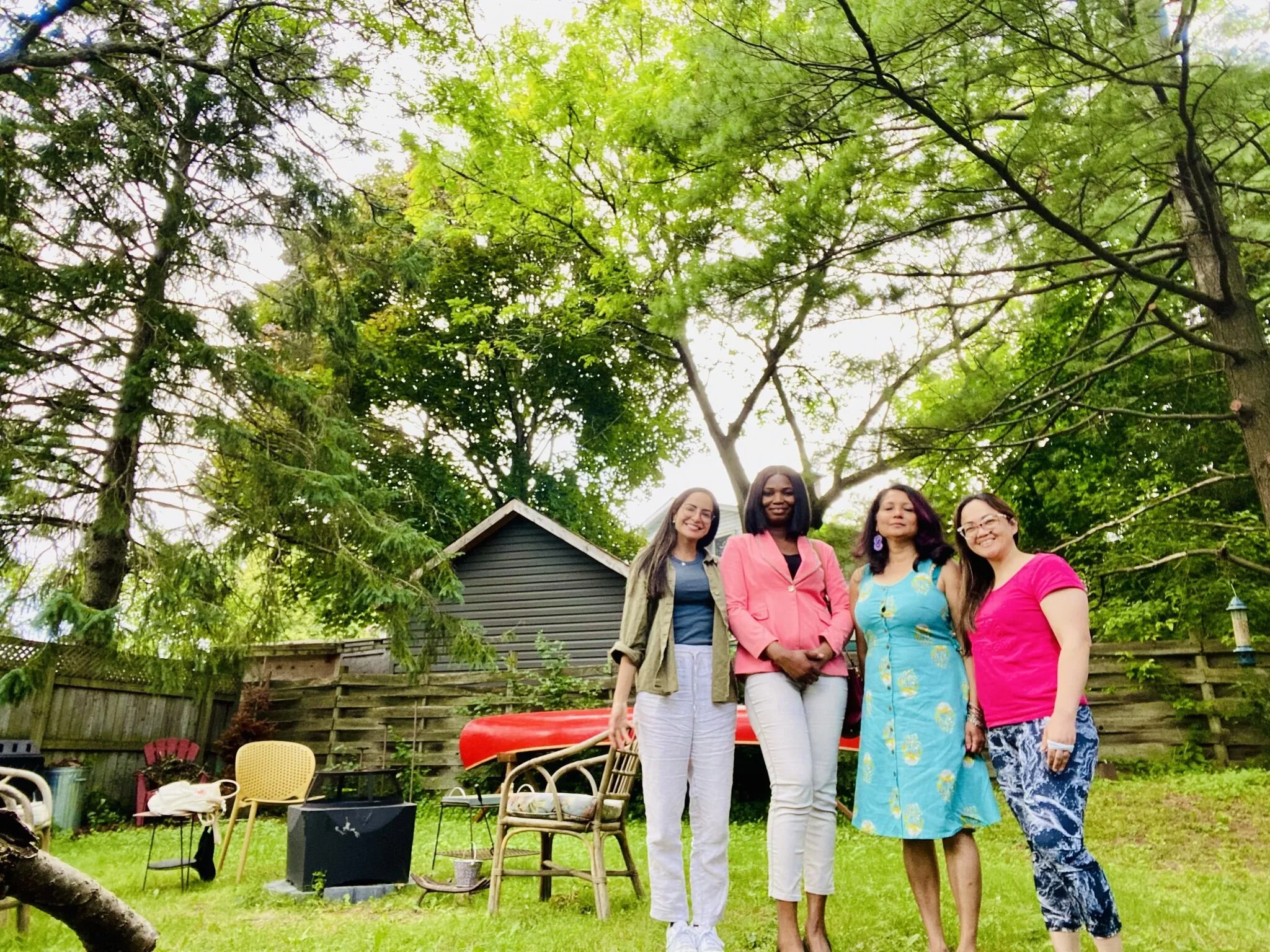 Four women standing outdoors in a backyard with trees and a fence, smiling, with chairs, a table, and a red canoe in the background.