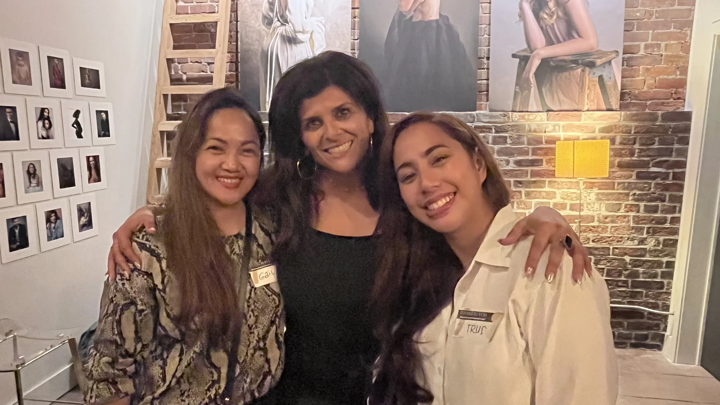 Three women smiling and posing together indoors, with artwork and photographs on brick and white walls behind them.