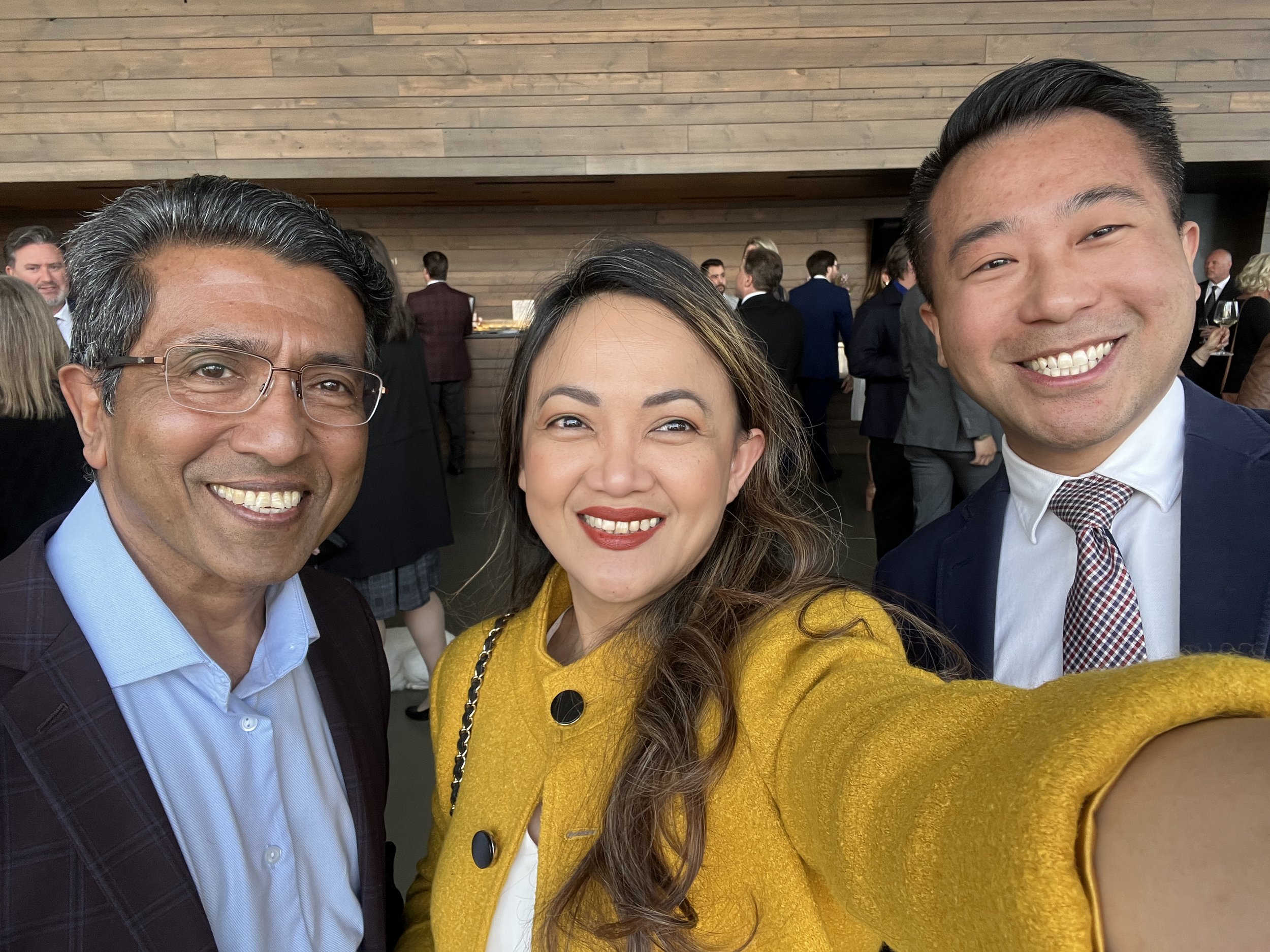 Three people taking a selfie at a formal event or gathering, with a wooden wall and other guests in the background.