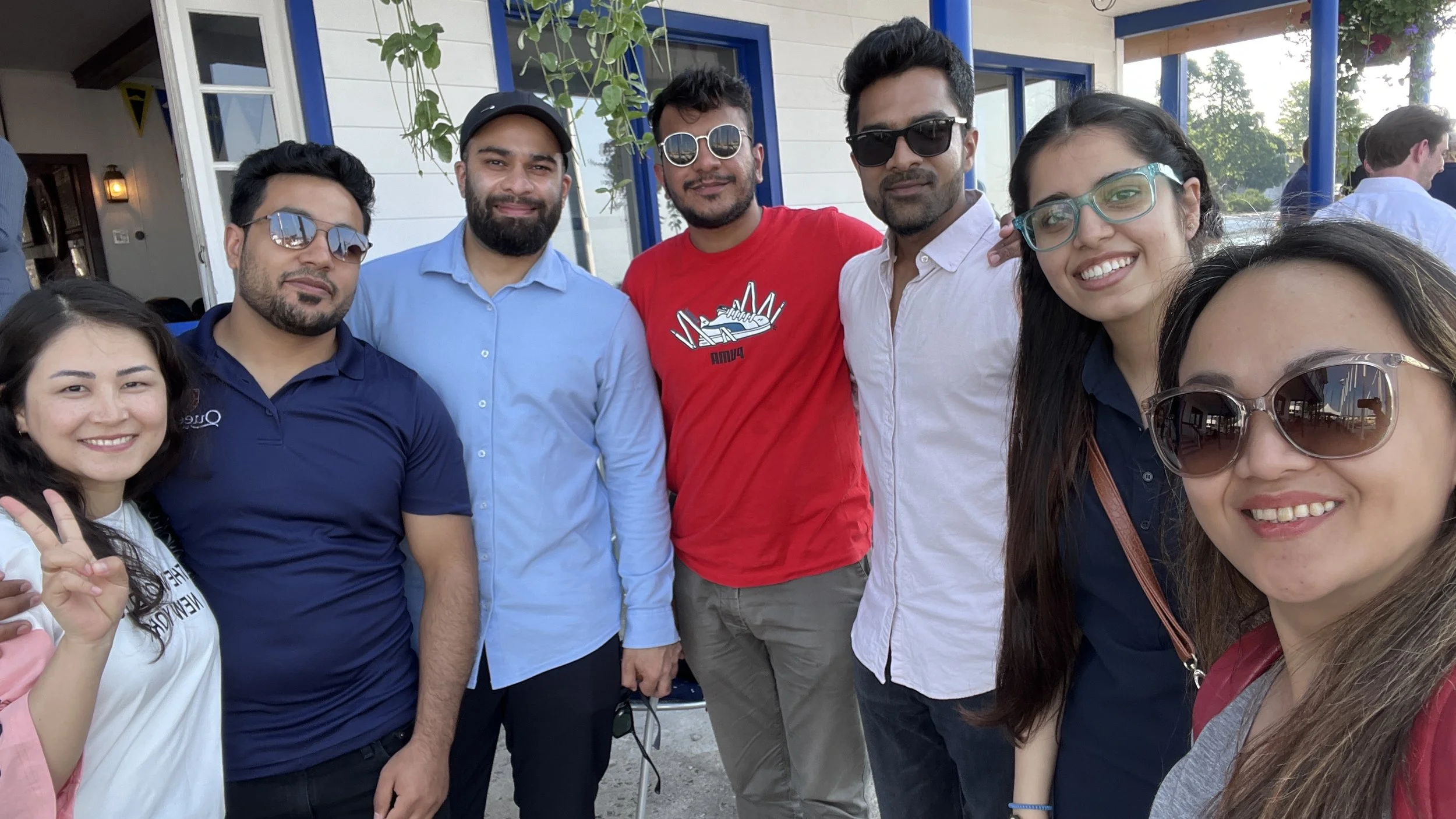 Group of seven young adults smiling outdoors, some wearing sunglasses, in front of a building with blue window frames, during daytime.