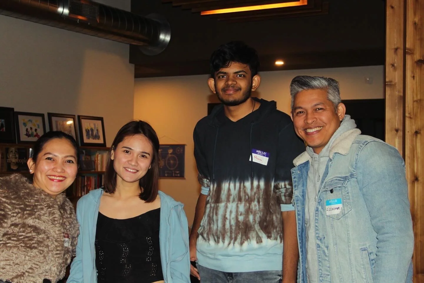 Group of four people smiling indoors, two women and two men, with framed pictures and shelves in the background.