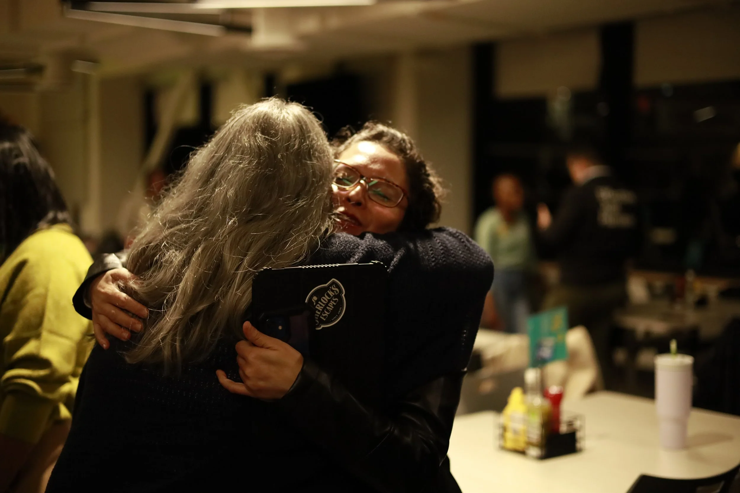 Two women hugging each other warmly in a social setting, with one woman smiling and wearing glasses, while the other has gray hair. The background shows other people and a table with condiments.