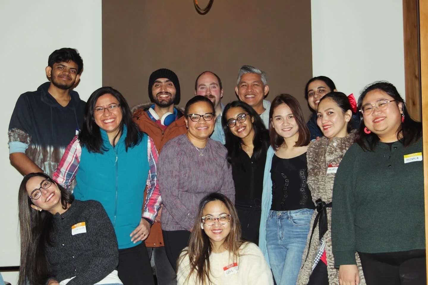 Group of diverse people smiling and posing together indoors, some with name tags, in a casual setting.
